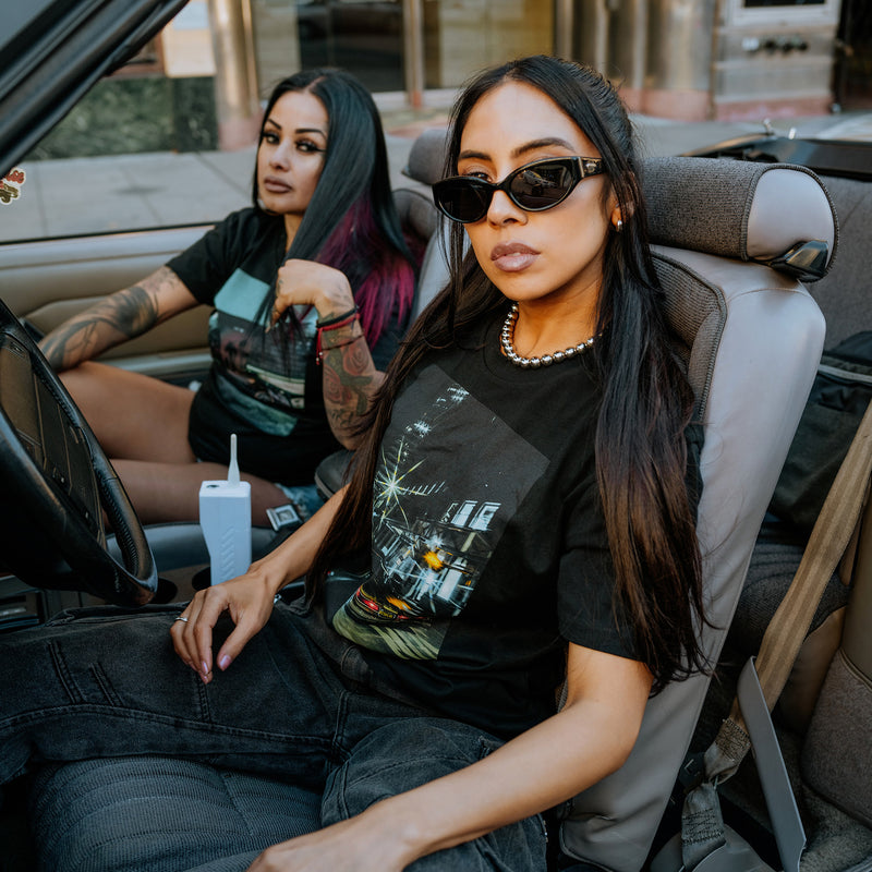 Two women sitting in a convertible vintage car wearing black Oaklandish t-shirts featuring Oakland Starry Night Cali Crusin image on the front chests.