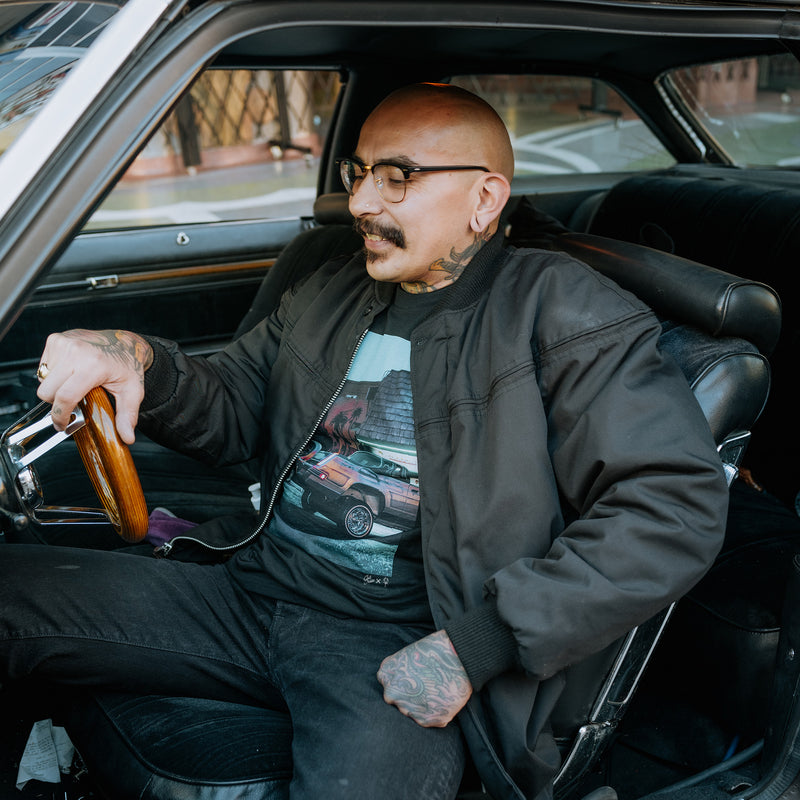 A man sitting in the driver's seat of a car with the door open wearing a black t-shirt with an image of Armando’s lowrider car parked in front of a Nipsey Hussle mural. 