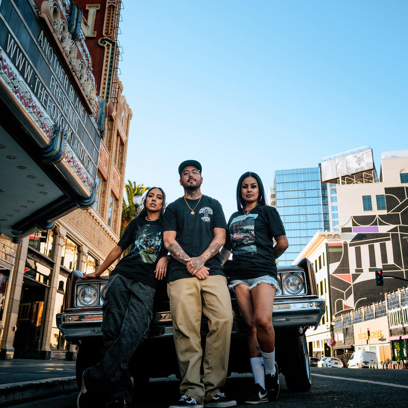 Two women and a man leaning against a vintage convertible in downtown Oakland wearing black Oaklandish t-shirts.