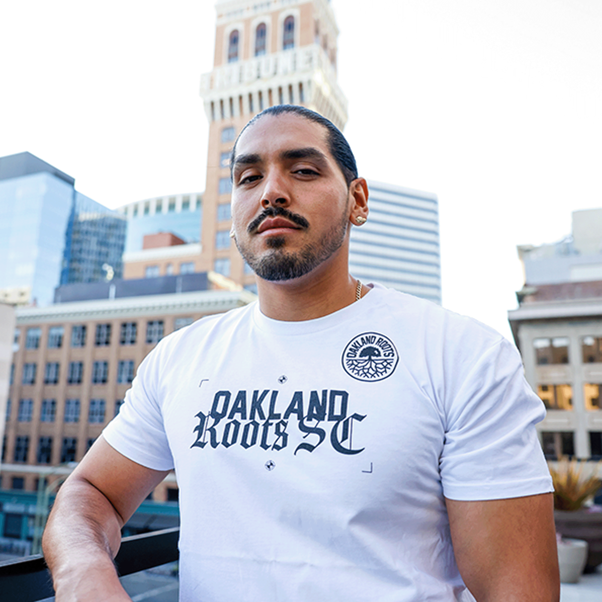 Man standing outdoors in Oakland wearing a white t-shirt with Oakland Roots SC wordmark with Old English script and black and white circle logo.