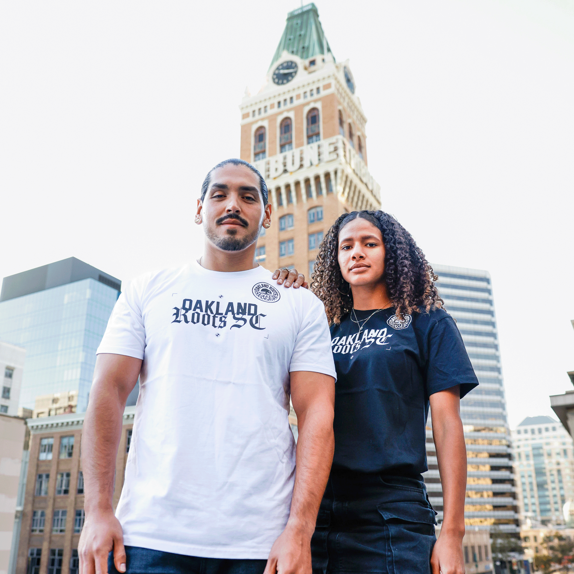 Man and woman are standing outdoors in Oakland wearing t-shirts with Oakland Roots RC wordmark and round logos. 