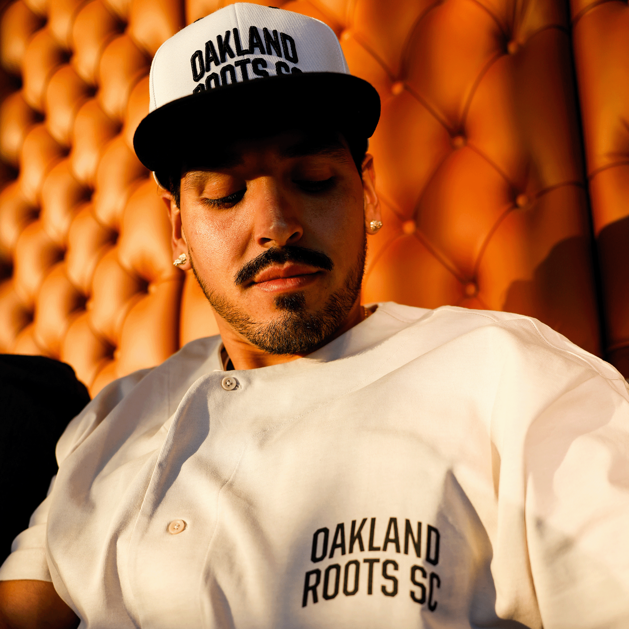 A person clad in an Oakland Roots SC Heavy Button-up Baseball Jersey and a white baseball cap, both proudly displaying the "Oakland Roots SC" brand, sits against an orange, tufted leather backdrop. With a mustache and goatee, they bask in the warm sunlight that illuminates the scene, perfectly embodying the social good ethos of the sports club.