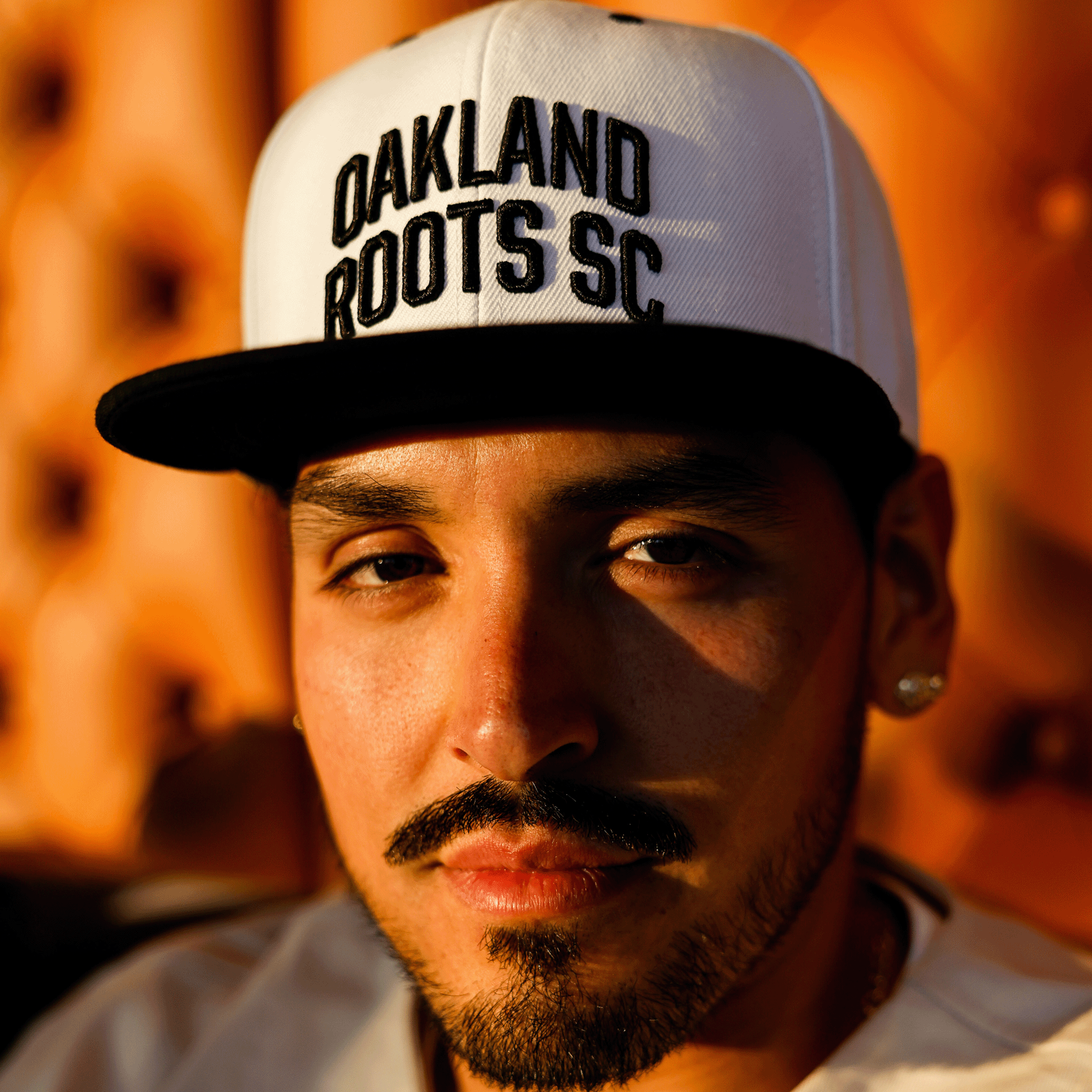 Close up of a man’s face wearing white snapback hat with black embroidered Oakland Roots SC wordmark on the crown and black flat square bill.