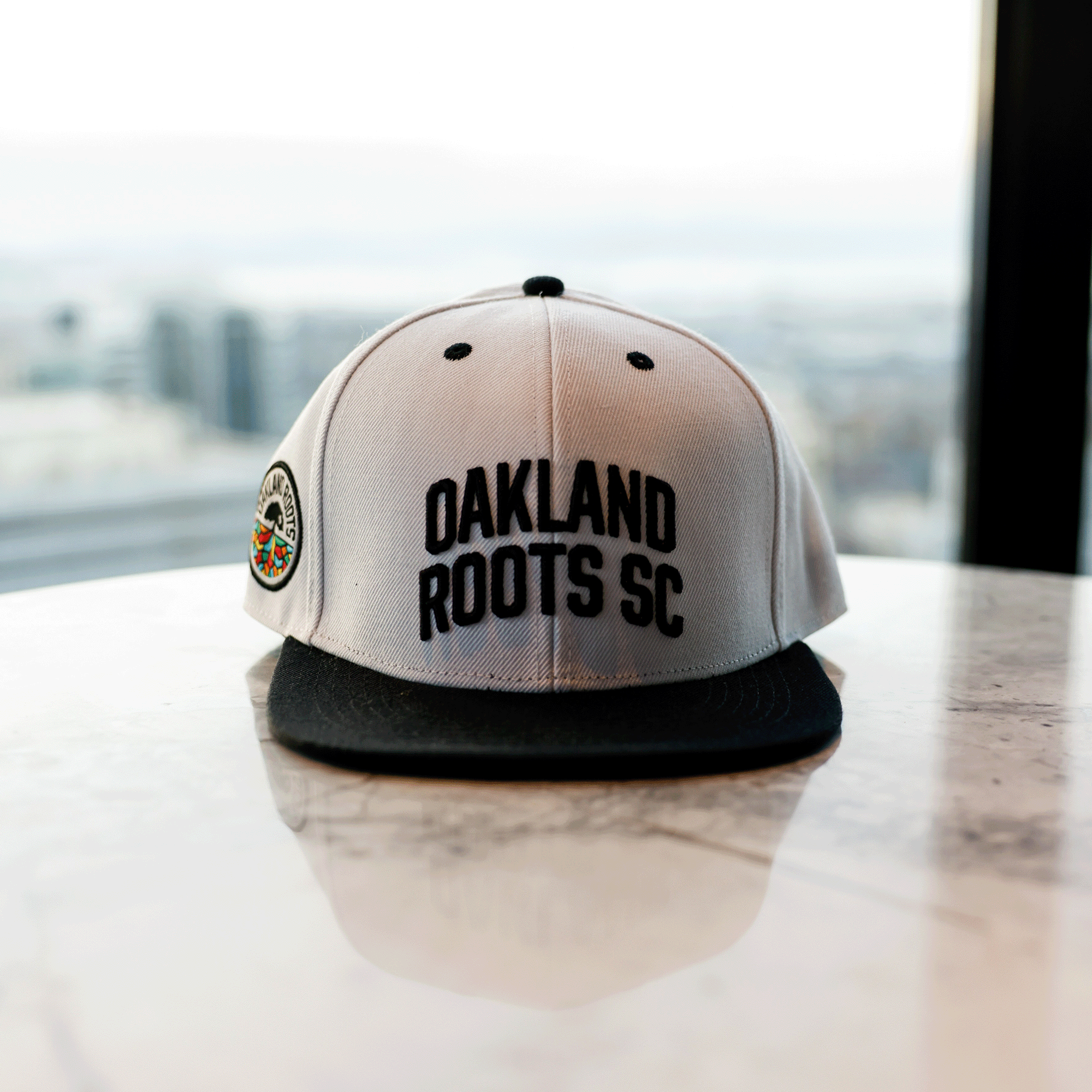White snapback hat with black embroidered Oakland Roots SC wordmark, full-color Roots SC logo, black flat square bill and black embroidered details sitting on counter top.