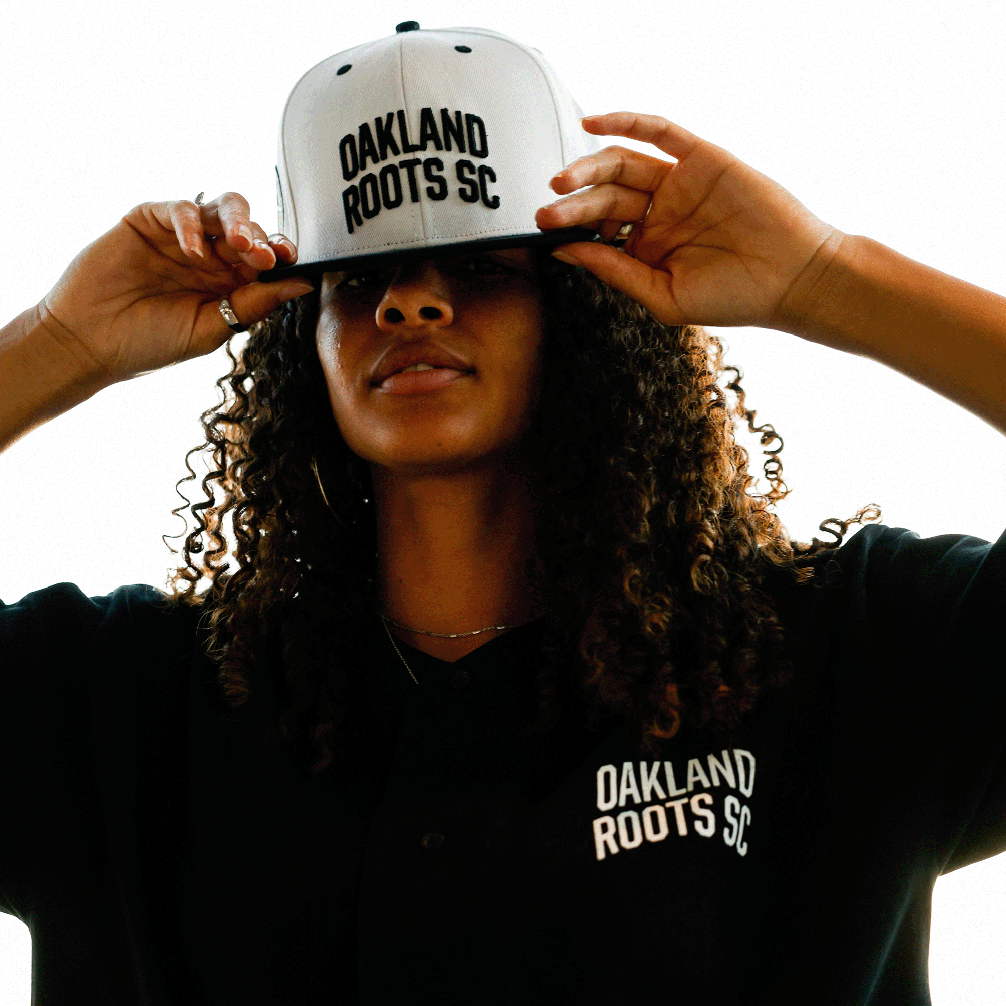 Woman wearing white hat with black embroidered Oakland Roots SC wordmark on the crown and black Oakland Roots SC t-shirt.