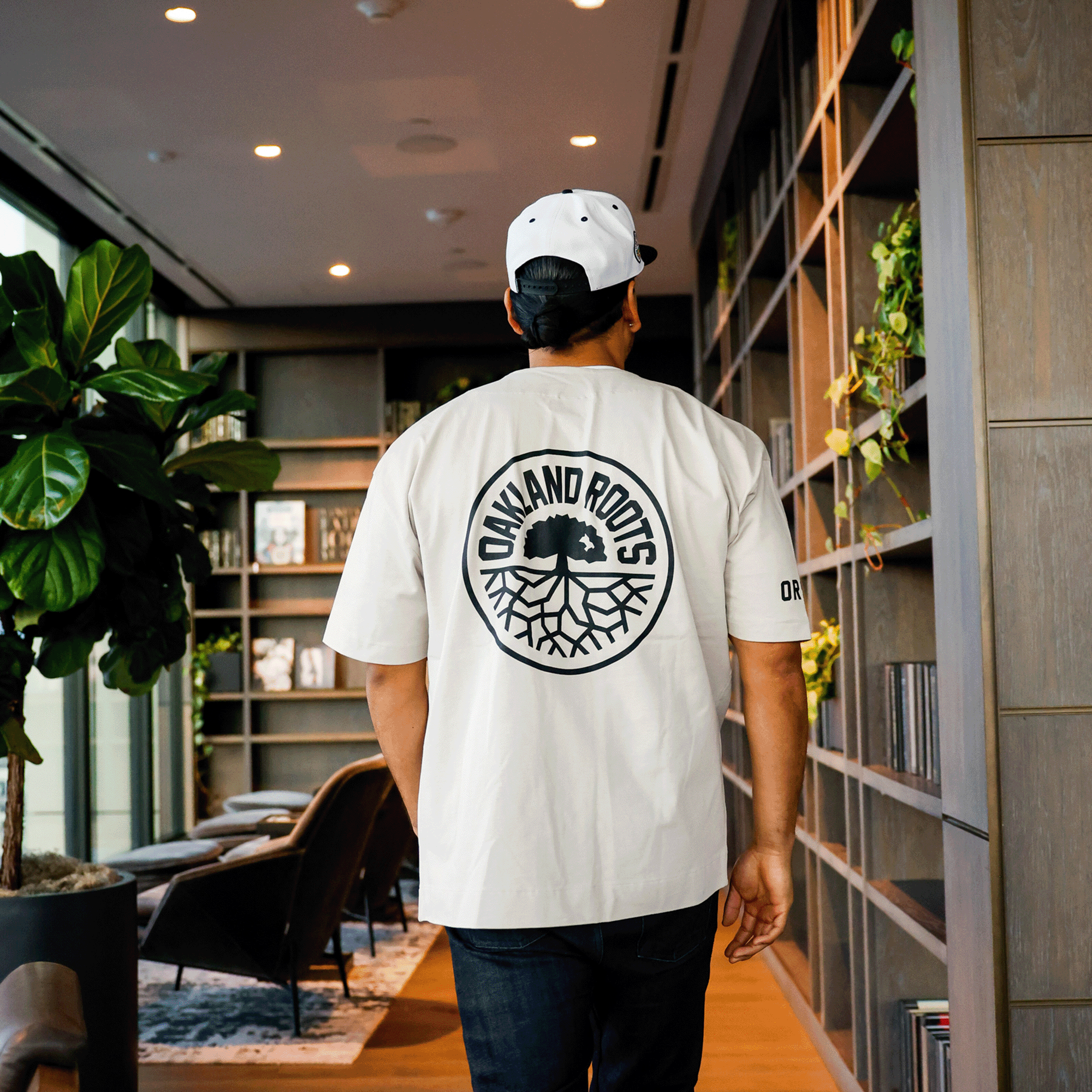 A person with short hair, dressed in an Oakland Roots SC Heavy Button-up Baseball Jersey and a white baseball cap, adorned with the "Oakland Roots" sports club and tree logo on the back, strolls through a well-lit room featuring wooden shelves filled with books and plants. In the background, a large plant and brown chairs arranged around a rug create an atmosphere of social good.