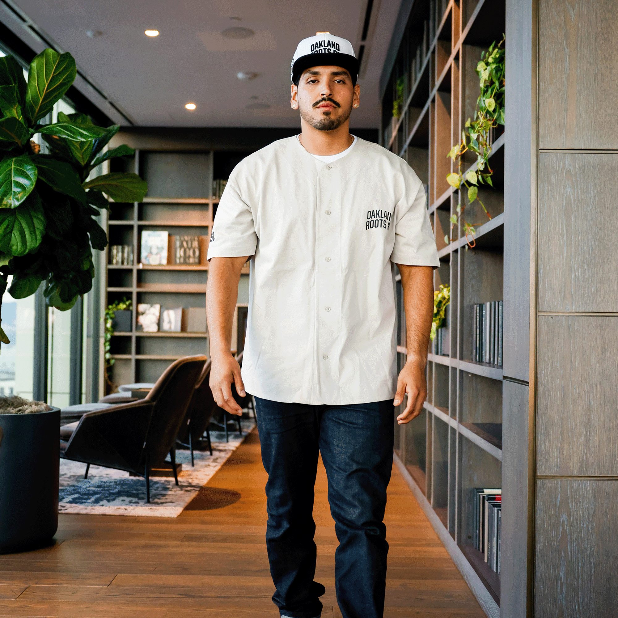 A man wearing an Oakland Roots SC Heavy Button-up Baseball Jersey and a matching cap stands inside a modern, stylish room with wooden floors, tall shelves filled with books, and potted plants. Representing the sports club known for its commitment to social good, he has a serious expression and his hands are relaxed at his sides.