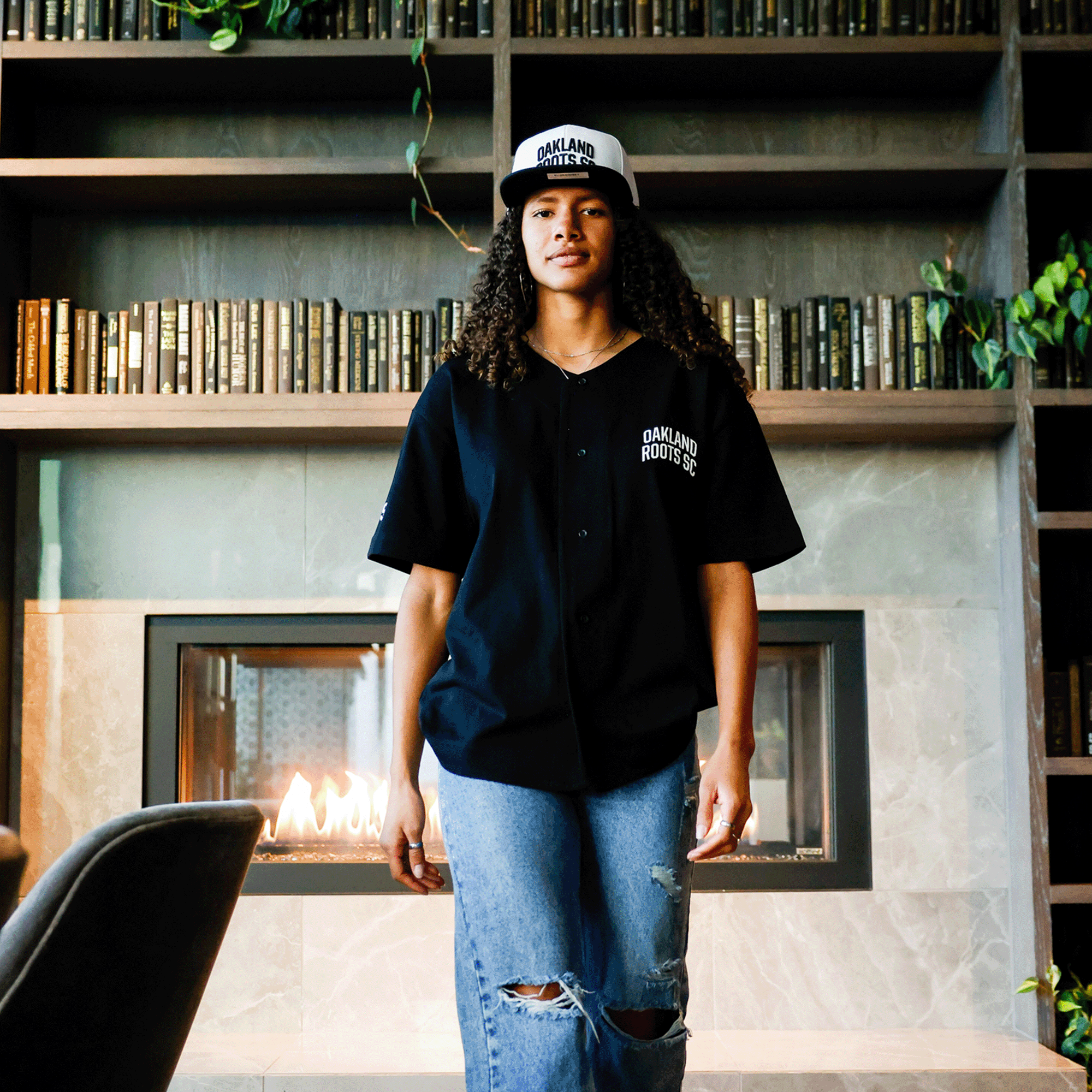 A person with curly hair, donning an Oakland Roots SC Heavy Button-up Baseball Jersey in black and an Oaklandish cap, stands in front of a modern fireplace. They're also sporting ripped jeans. Behind them, shelves laden with books and decorative plants add a cozy, stylish ambiance to the room.