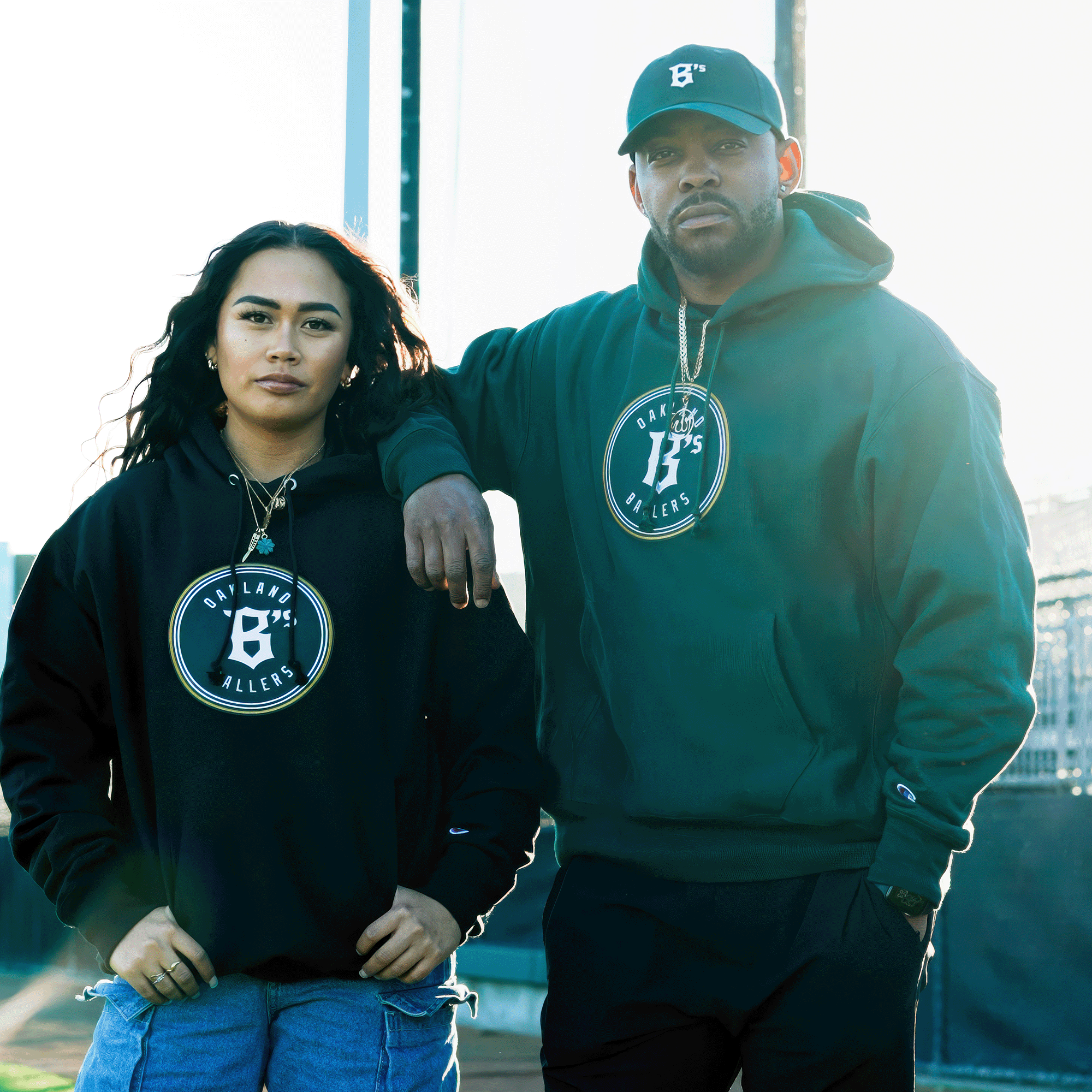 On a sunny day, two people confidently wear Champion X Oakland Ballers Logo Hoodies featuring the "B" logo and "Bay Lane Millers" text. The person on the right pairs it with a matching cap, posing in front of a blurred athletic field, representing the Oakland Ballers spirit.