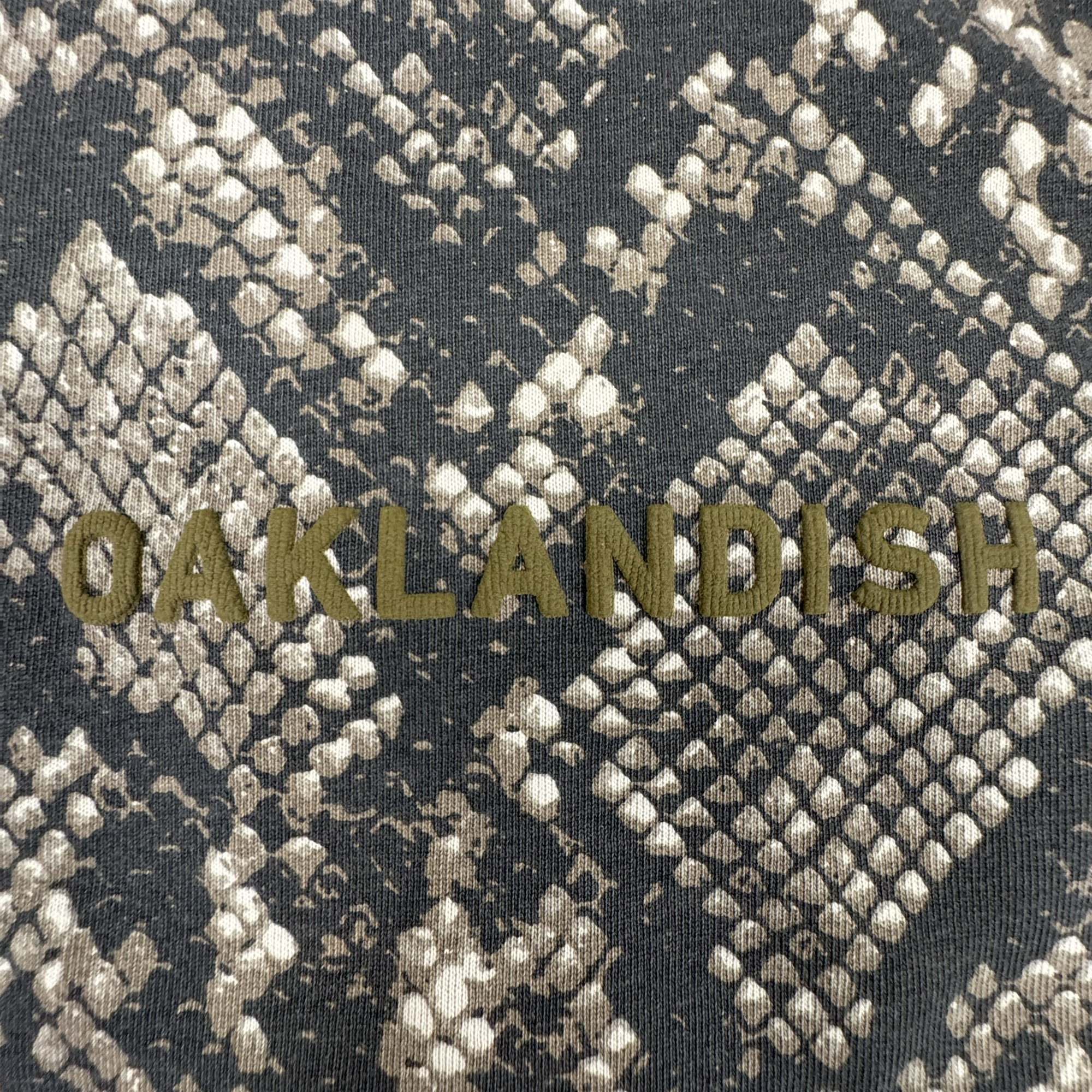 Crafted from 100% cotton for both comfort and style, the Oaklandish Classic Logo LS Tee by Oaklandish features a textured gray and tan snakeskin patterned background. Centered on the long sleeve t-shirt are bold, olive-green “OAKLANDISH” letters in an all-caps, sans-serif font. The design is complemented by vintage black sleeves for a unique look.