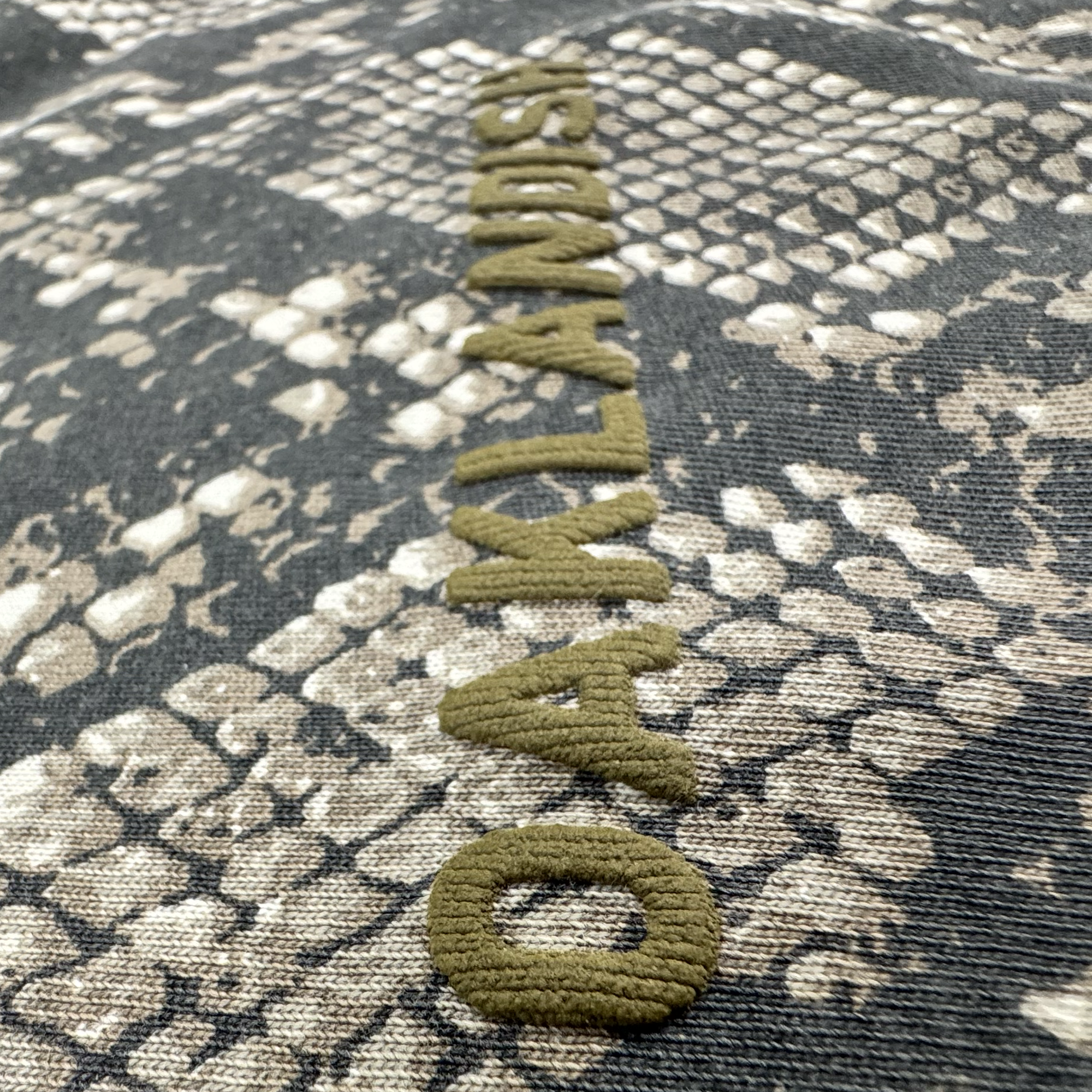 Close-up of the fabric on the Oaklandish Classic Logo LS Tee by Oaklandish, featuring a textured brown and beige snakeskin print on a black long sleeve 100% cotton shirt. The classic Oaklandish logo is stitched vertically in raised olive-green thread, contrasting against the reptile-inspired pattern and highlighting the detailed embroidery and textile quality.