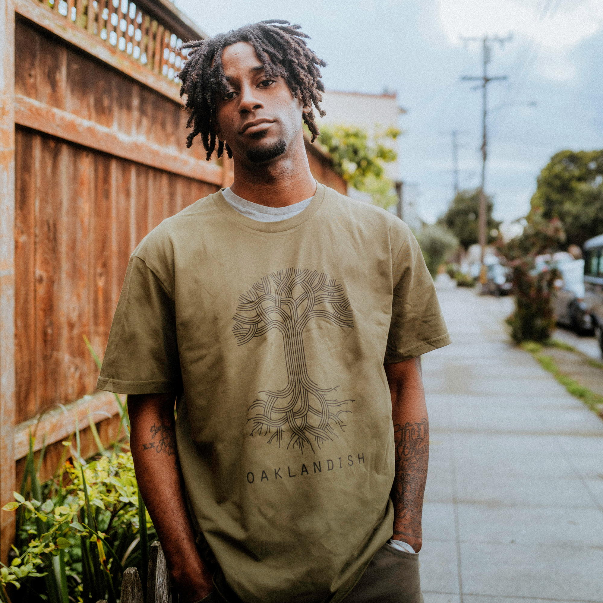 A person stands on a sidewalk wearing the olive green Oaklandish Classic Logo Tee with a tree design and "OAKLANDISH" on 100% cotton. They have short dreadlocks, looking at the camera, framed by a wooden fence and greenery, with houses and trees in the backdrop.