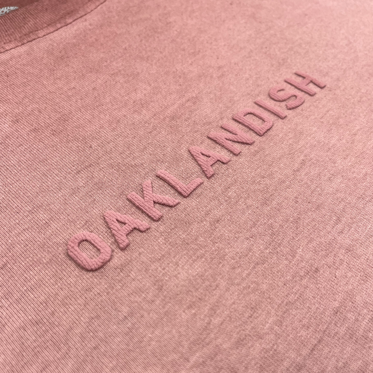 A close-up of the Oaklandish Classic Logo Tee by Oaklandish, showcasing heavyweight pink cotton fabric with “OAKLANDISH” embroidered in raised, matching pink letters across the chest. The detailed texture and stitching emphasize its quality and classic men’s sizing.