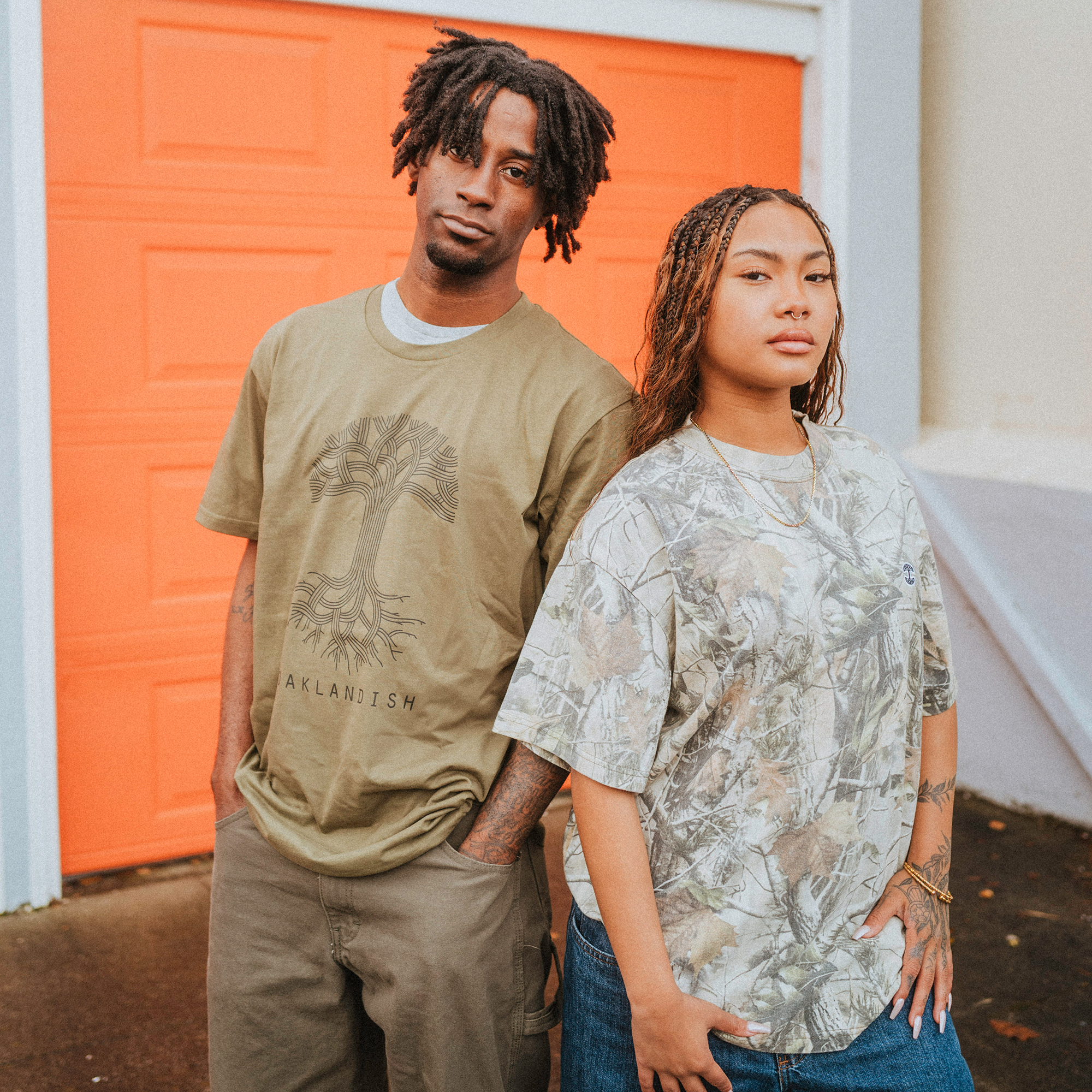 With dreadlocks, one person sports an olive green Oaklandish Classic Logo Tee by Oaklandish, standing next to a second individual with long braids in a camo-patterned shirt. They’re in front of an orange garage door on concrete, both displaying neutral expressions that convey a laid-back vibe.