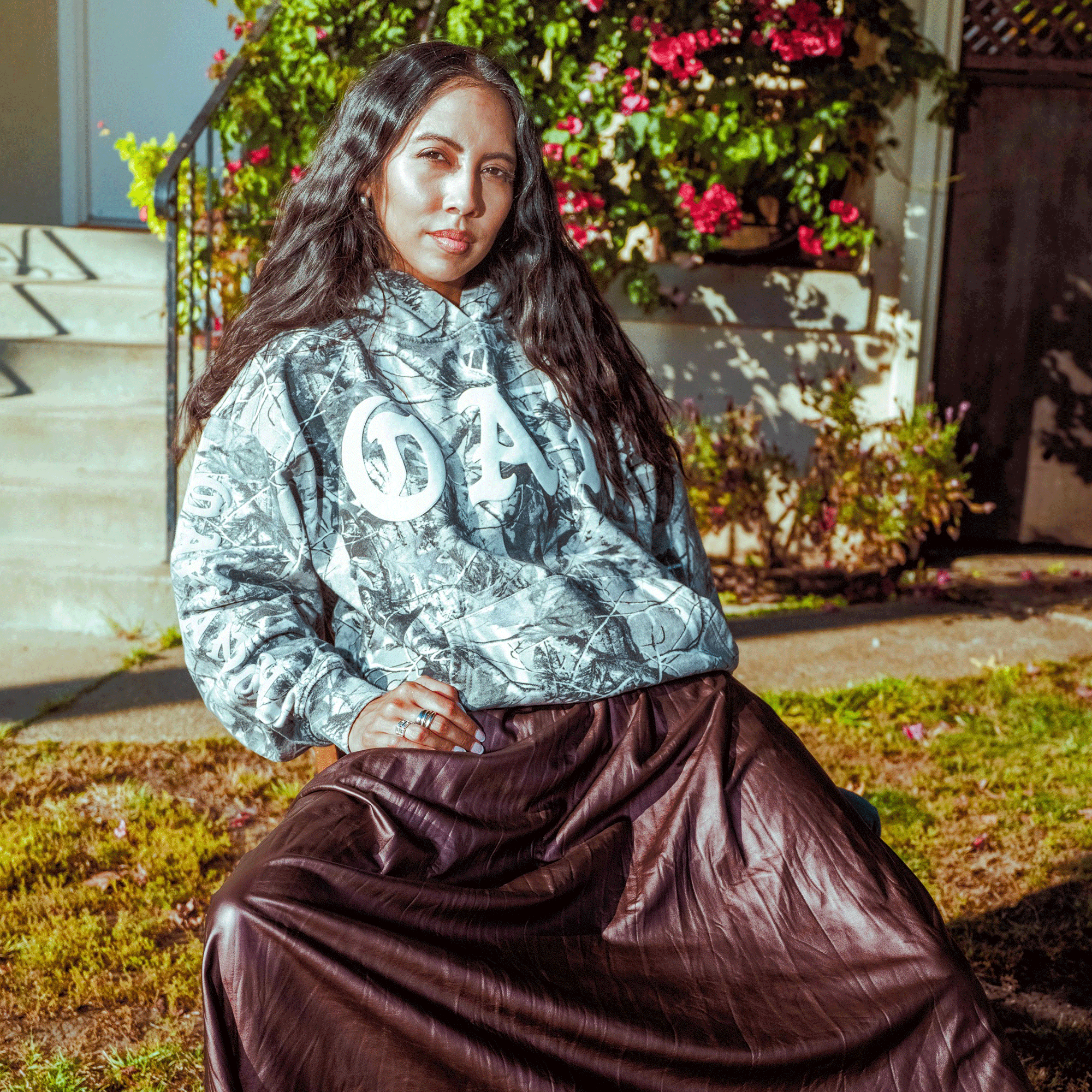 A woman with long dark hair sits outside on a sunny day, wearing the Oaklandish Cork Hoodie and a long brown skirt. She smiles slightly, surrounded by flowers, greenery, and house steps in the background.