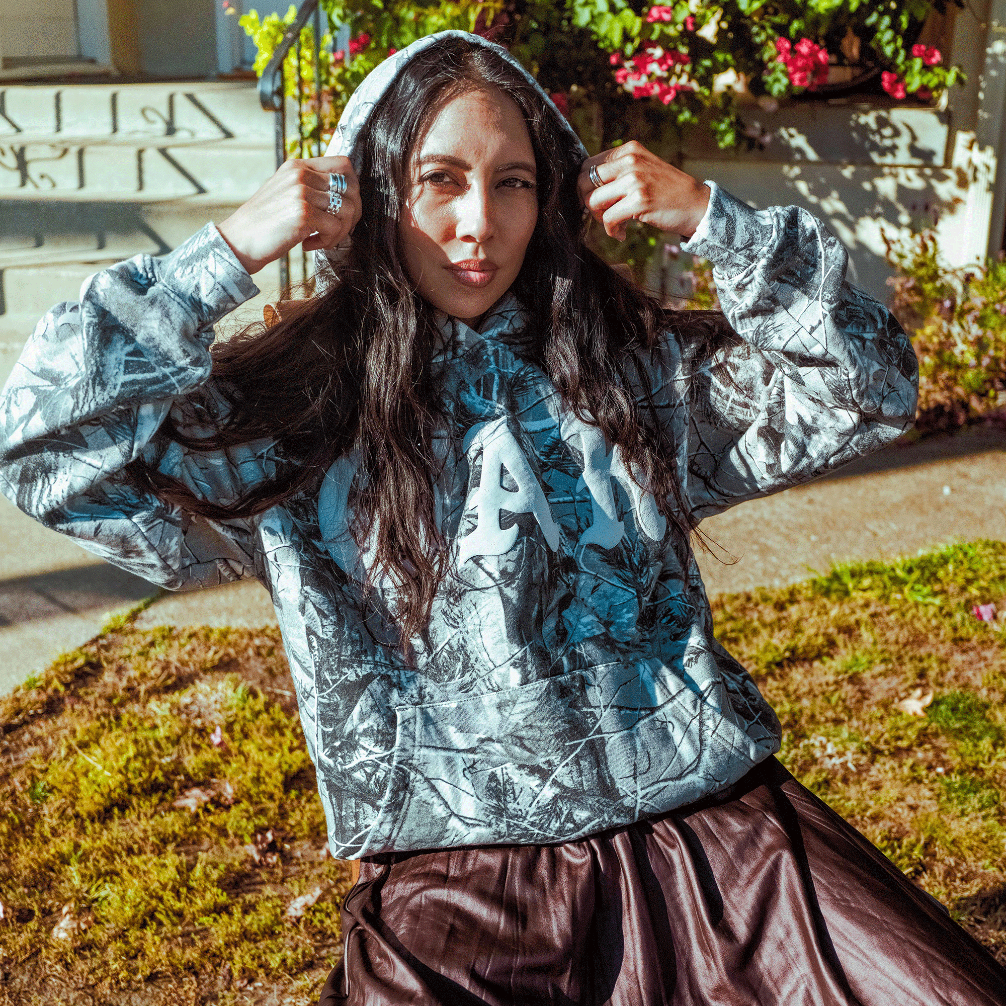 A woman with long dark hair sits outside on a sunny day, pulling up the hood of her Oaklandish Cork Hoodie, which features a grey wood camo pattern and bold white letters. She pairs it with a brown skirt and multiple rings, surrounded by flowers and greenery.