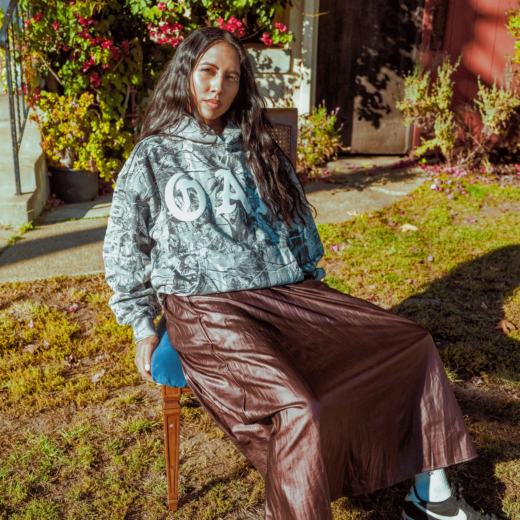 A woman with long dark hair sits outdoors on a chair, enjoying the sun in a Cork Hoodie by Oaklandish and a brown skirt, surrounded by greenery and flowers in a relaxed, casual setting.