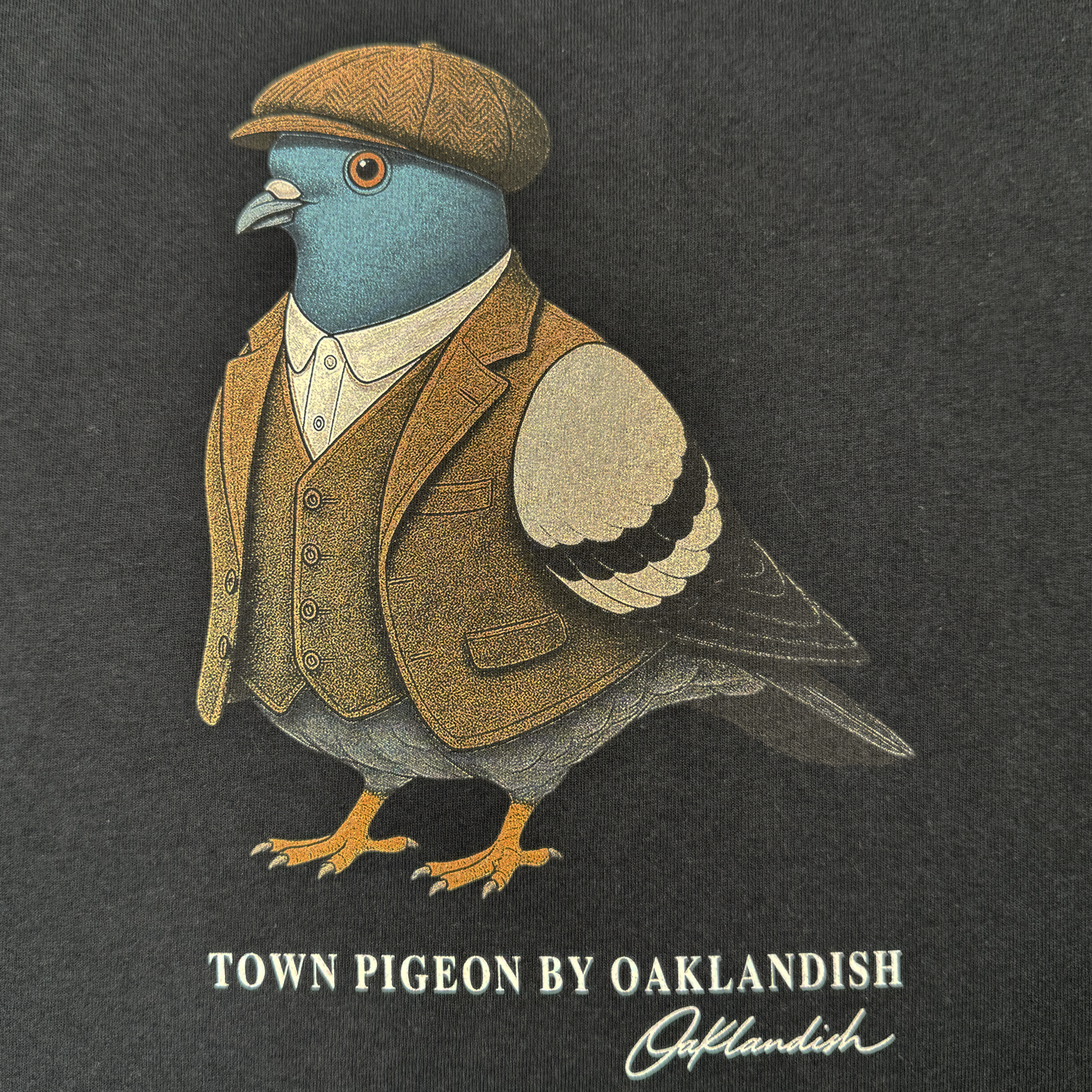 A pigeon sporting a brown tweed suit, vest, and flat cap stands upright against a dark background, capturing the timeless style of the men’s collection. Below it reads “Dapper Tee by Oaklandish” with the Oaklandish logo signature underneath—ideal for a classic fit t-shirt in heavyweight cotton.