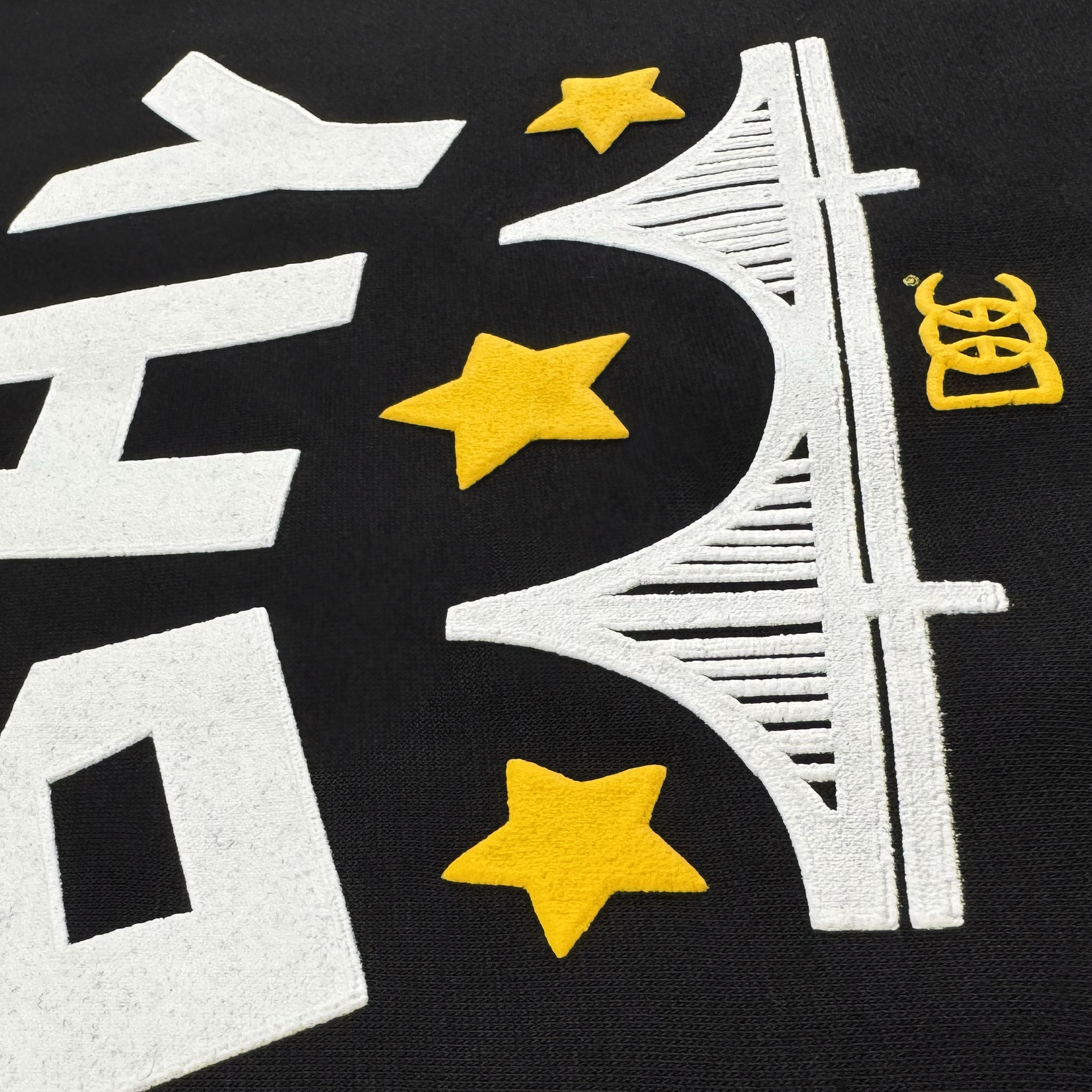 The Bay Bridge Hoodie By DOC V2 from Oaklandish features a black background with a white stylized bridge, three yellow stars, bold geometric shapes, and a small yellow symbol—embroidered or printed as part of The Bay Collection.