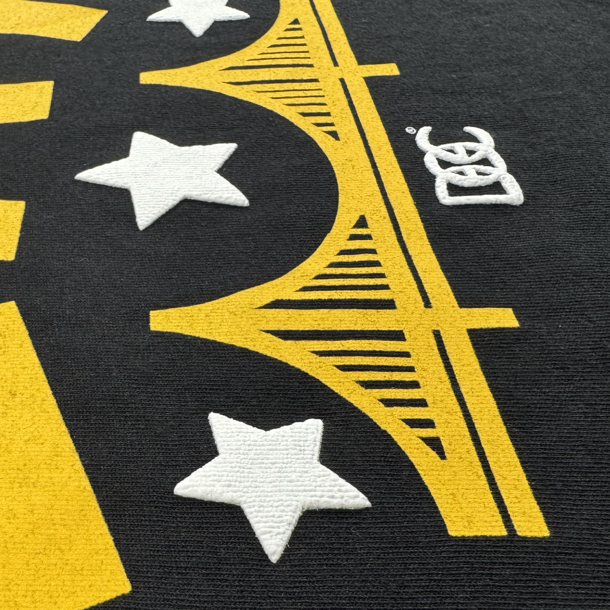 A close-up of the Oaklandish Bay Bridge Tee By DOC V2 shows its heavyweight cotton, bold yellow bridge design, three white embroidered stars, a small white interlocking rings logo, and detailed stitching.