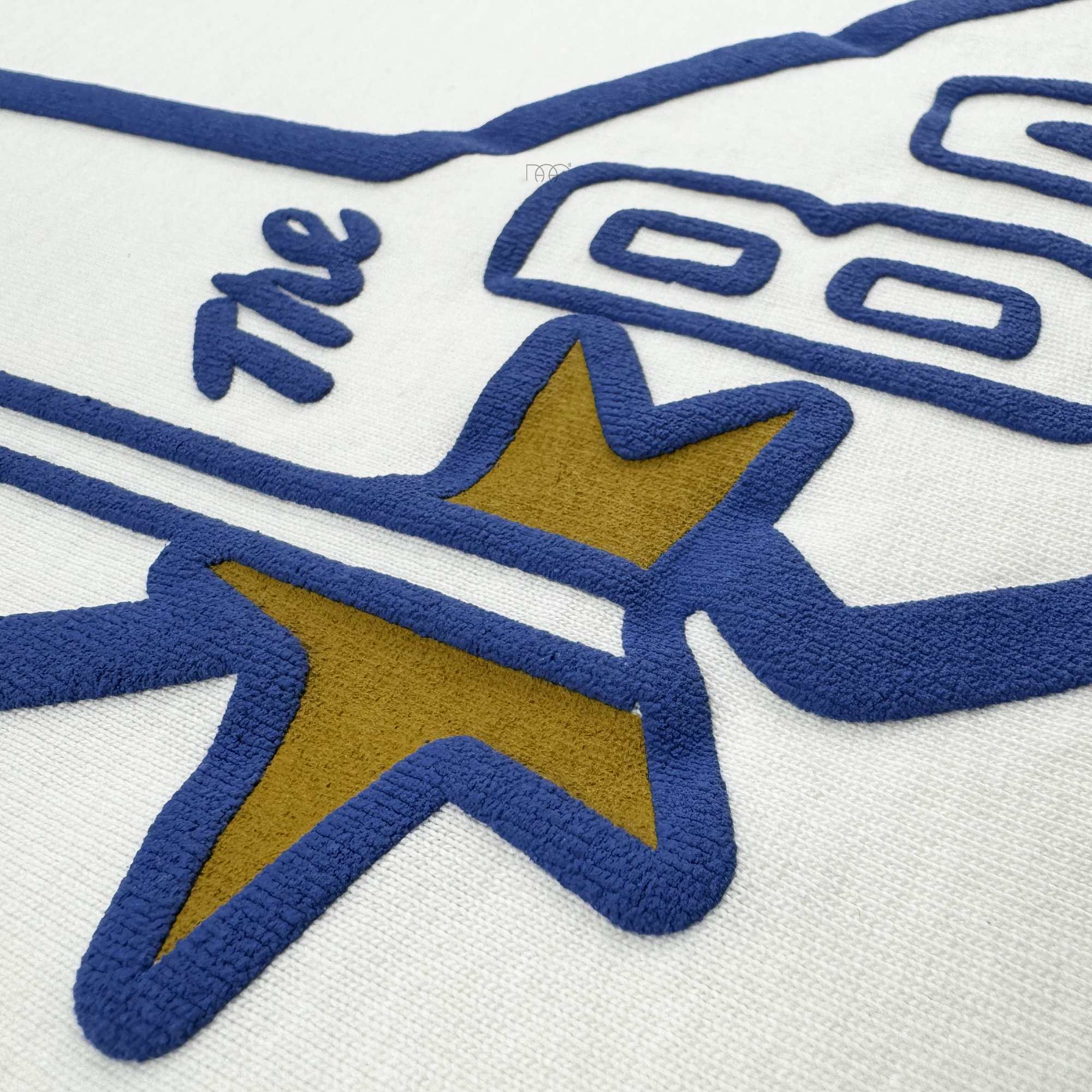 A close-up of heavyweight cotton on the CA Bay Area Tee By DOC from Oaklandish shows blue and gold embroidery outlining a stylized airplane, with teal cursive "The" above the wing—part of The Bay Collection.