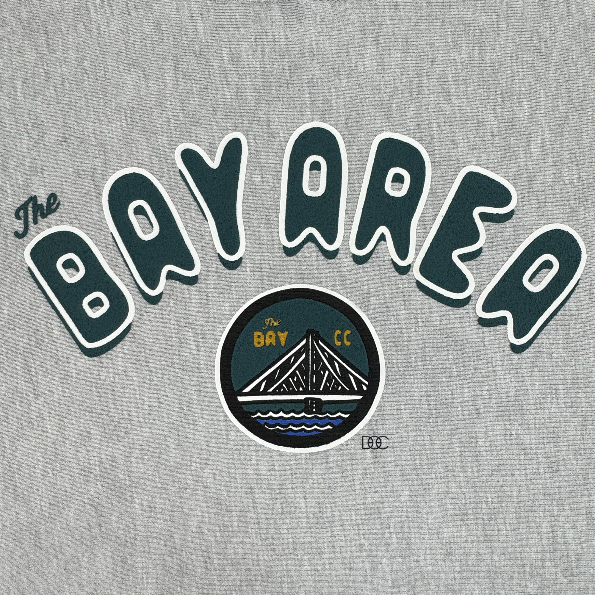 The Bay Area CC Hoodie by DOC from Oaklandish is a gray men’s pullover featuring “The Bay Area” in dark green outlined in white and a circular emblem with a bridge, “The Bay,” and “CC” in yellow for San Francisco Bay Area pride.