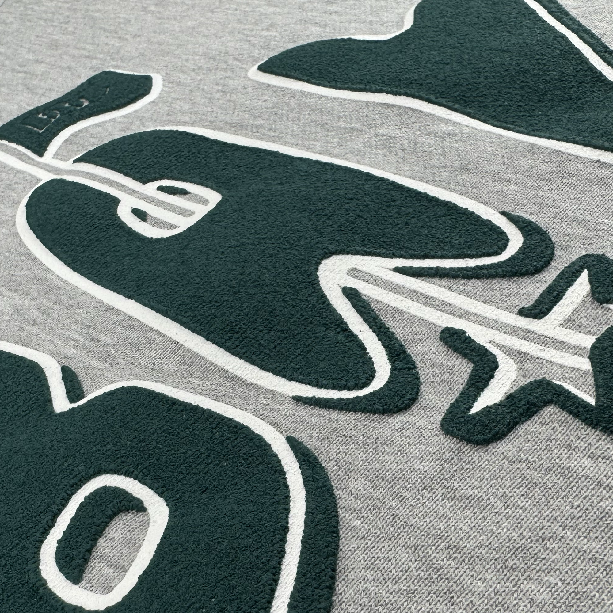 Close-up of textured dark green appliqué letters and a star on light gray premium heavyweight crewneck fleece from the Oaklandish Bay Flag Crew by DOC. The white-outlined "G" has visible stitching and a small flag atop it.