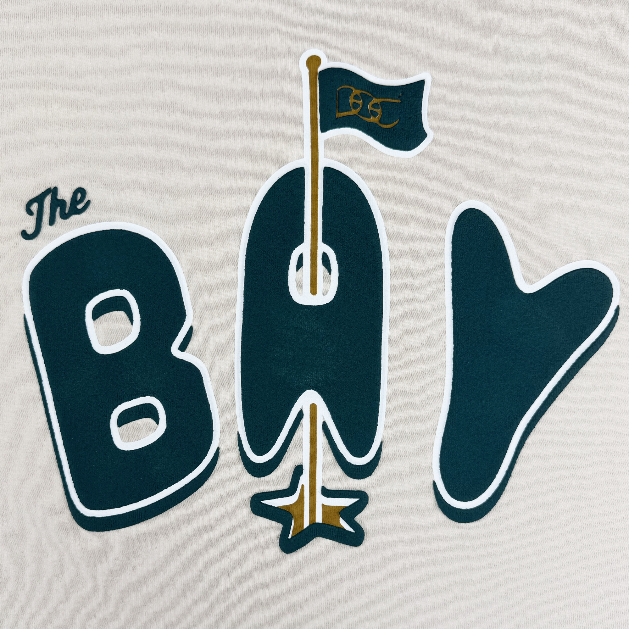 The image showcases "The BAY" in bold dark green, with the "A" as a flagpole topped by a gold star and flying a "BSC" flag. Inspired by The Bay Collection, this design appears on the heavyweight cotton Bay Flag Tee By DOC from Oaklandish.