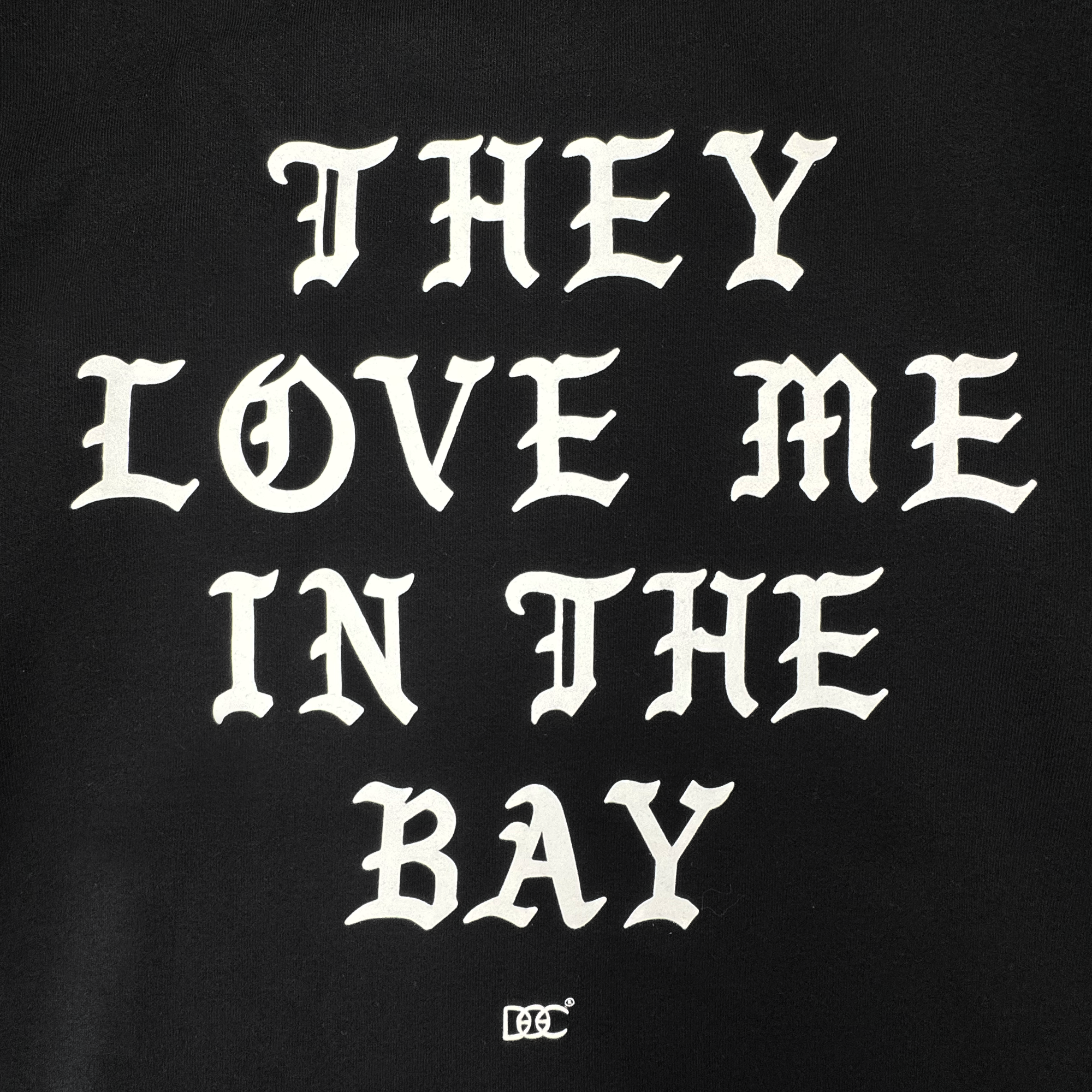 White Gothic-style text on a black background reads: “THEY LOVE ME IN THE BAY.” Centered beneath the statement, a small decorative By DOC logo enhances the classic streetwear aesthetic. Crafted as part of The Bay Collection, the They Love Me Zip By Doc from By DOC delivers iconic style to your wardrobe—perfect for layering with your favorite heavyweight fleece or zip-up hoodie.