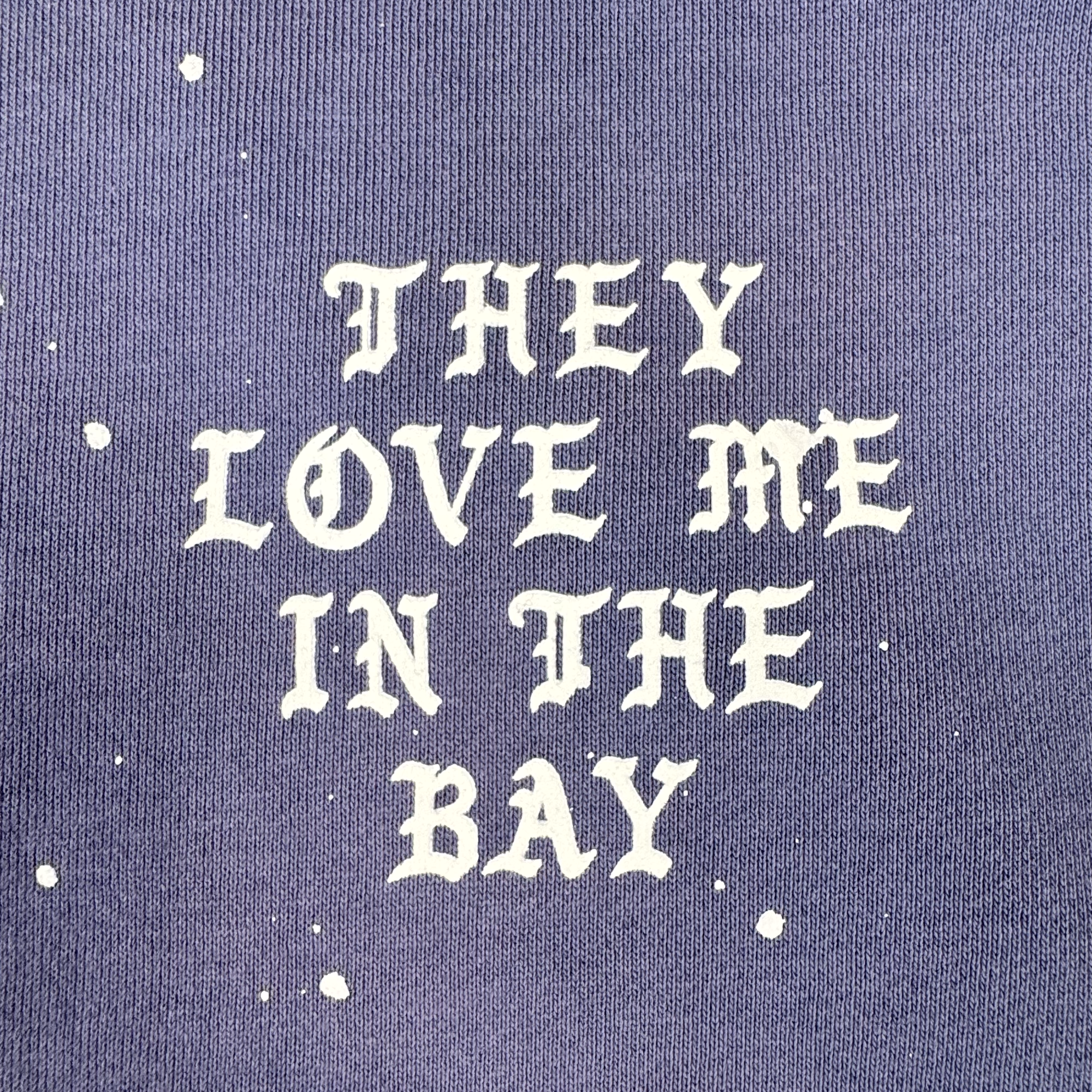 White gothic-style text reading “THEY LOVE ME IN THE BAY” stands out against purple fabric, accented with random white paint splatters. This bold design turns heads and adds a unique flair to any wardrobe—especially when featured on the They Love Me Hoodie By Doc by BY DOC, a heavyweight fleece men’s pullover hoodie designed for both comfort and style.