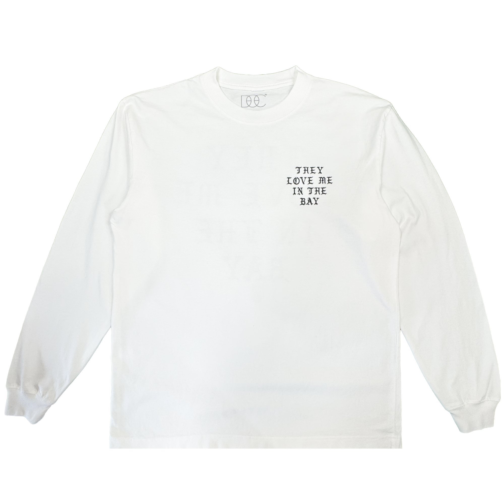 A heavyweight cotton long sleeve t-shirt by By DOC, the They Love Me LS Tee By Doc, is shown in men’s sizing laid flat against a plain white background. The only visible design is small black text on the left chest that reads, “THEY LOVE ME IN THE BAY.” There are no other embellishments or graphics.