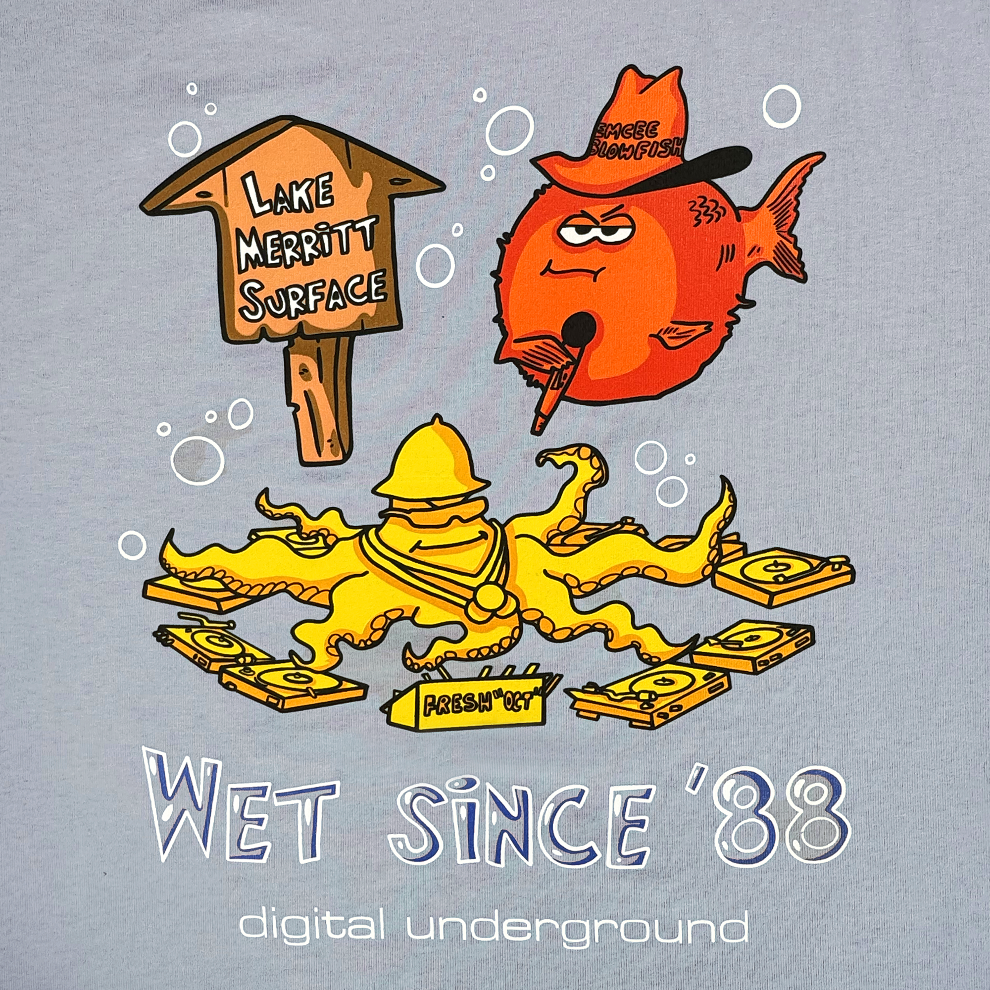A cartoon on the Oaklandish Wet Since '88 Heavy Tee features an orange fish with Oakland roots, wearing a cowboy hat, smoking a cigar by a "Lake Merritt Surface" sign. Nearby, an octopus holds cassettes and records. Text reads "Wet since '88" and "Underwater Rimes" by Digital Underground.
