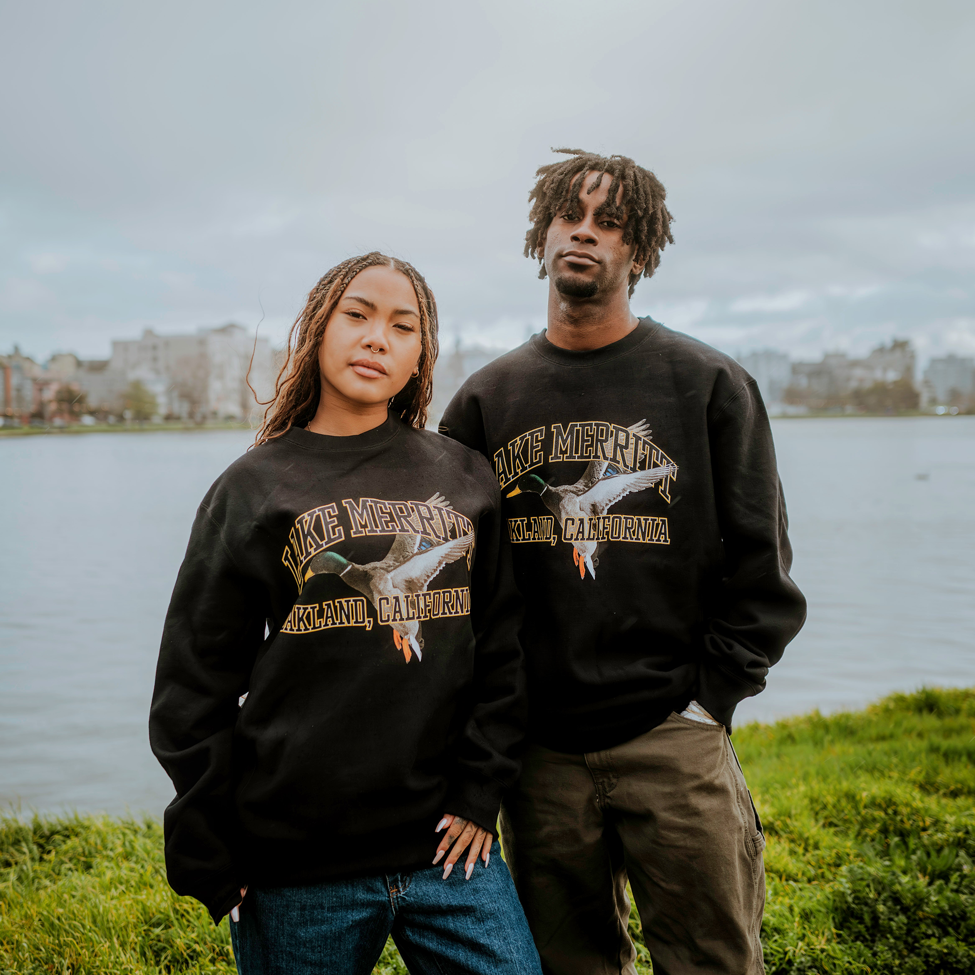 A man and woman stand on the grass by a lake, both wearing Oaklandish's Duck School Heavy Crew sweatshirts featuring "Lake Merritt Oakland, California" and an eagle graphic. Cloudy skies and distant buildings form the backdrop. The woman has braided hair; the man has short dreadlocks.