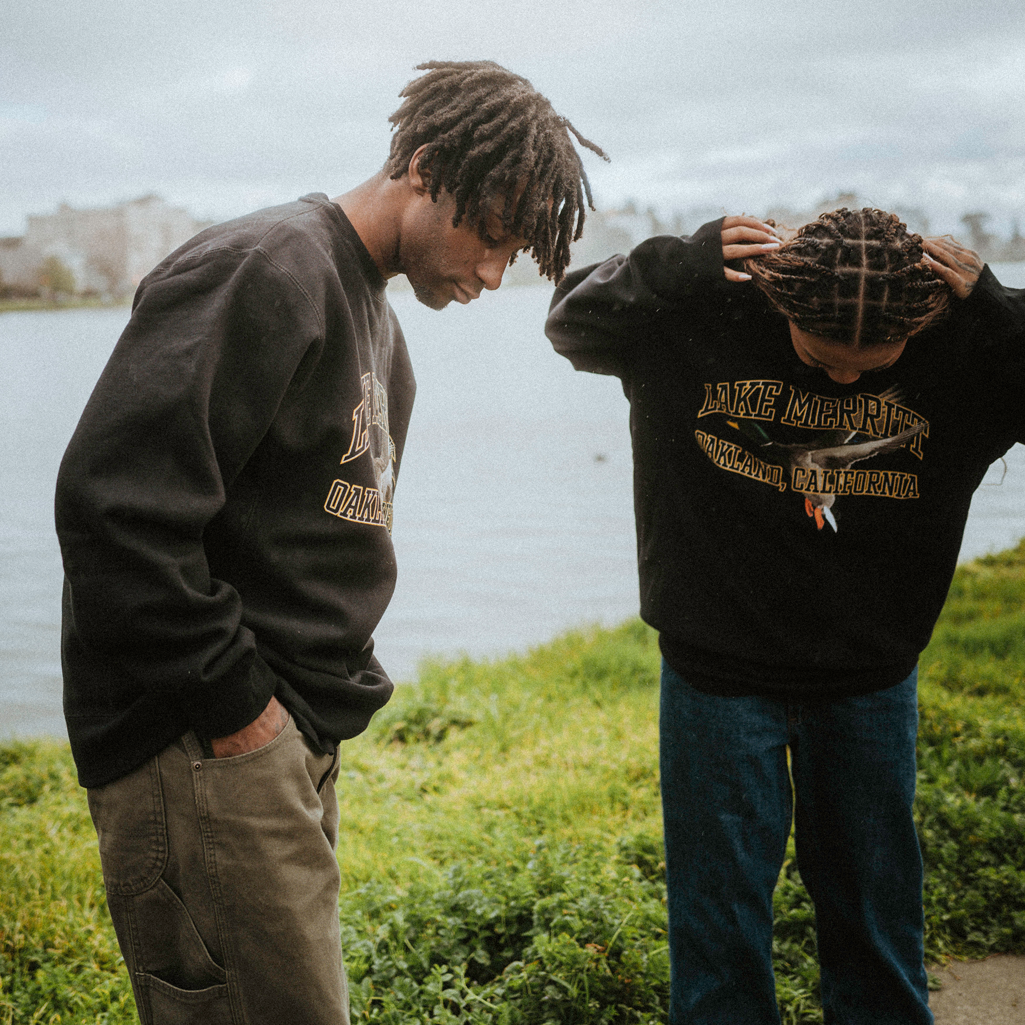 Two people stand by a lakeside wearing dark Oaklandish Duck School Heavy Crew sweatshirts, featuring "Lake Merritt, Oakland, California." The person on the left has short dreadlocks and looks down, while the person on the right adjusts their long braided hair. Water and greenery in the background.