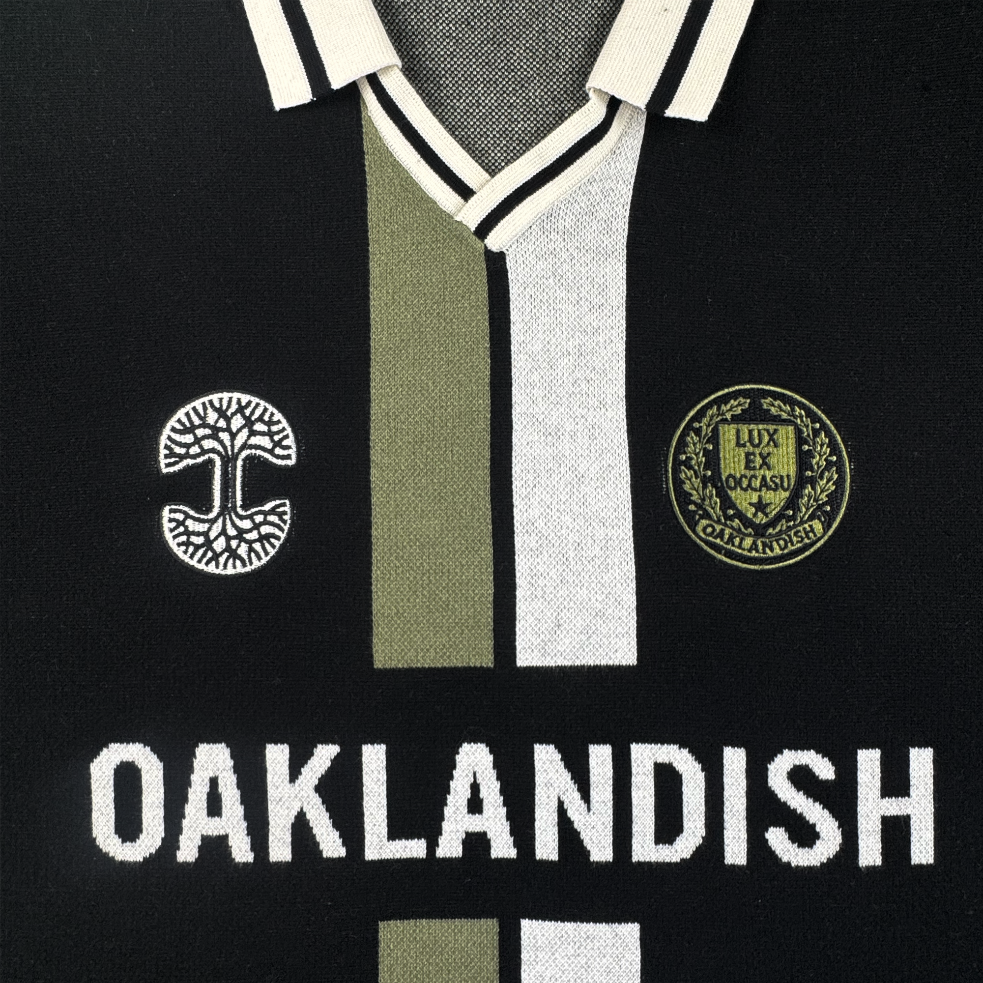 The Oaklandish Ease Jersey is a unisex black knit-collar top with white and olive-green stripes, two embroidered patches, and bold "OAKLANDISH" lettering across the chest.