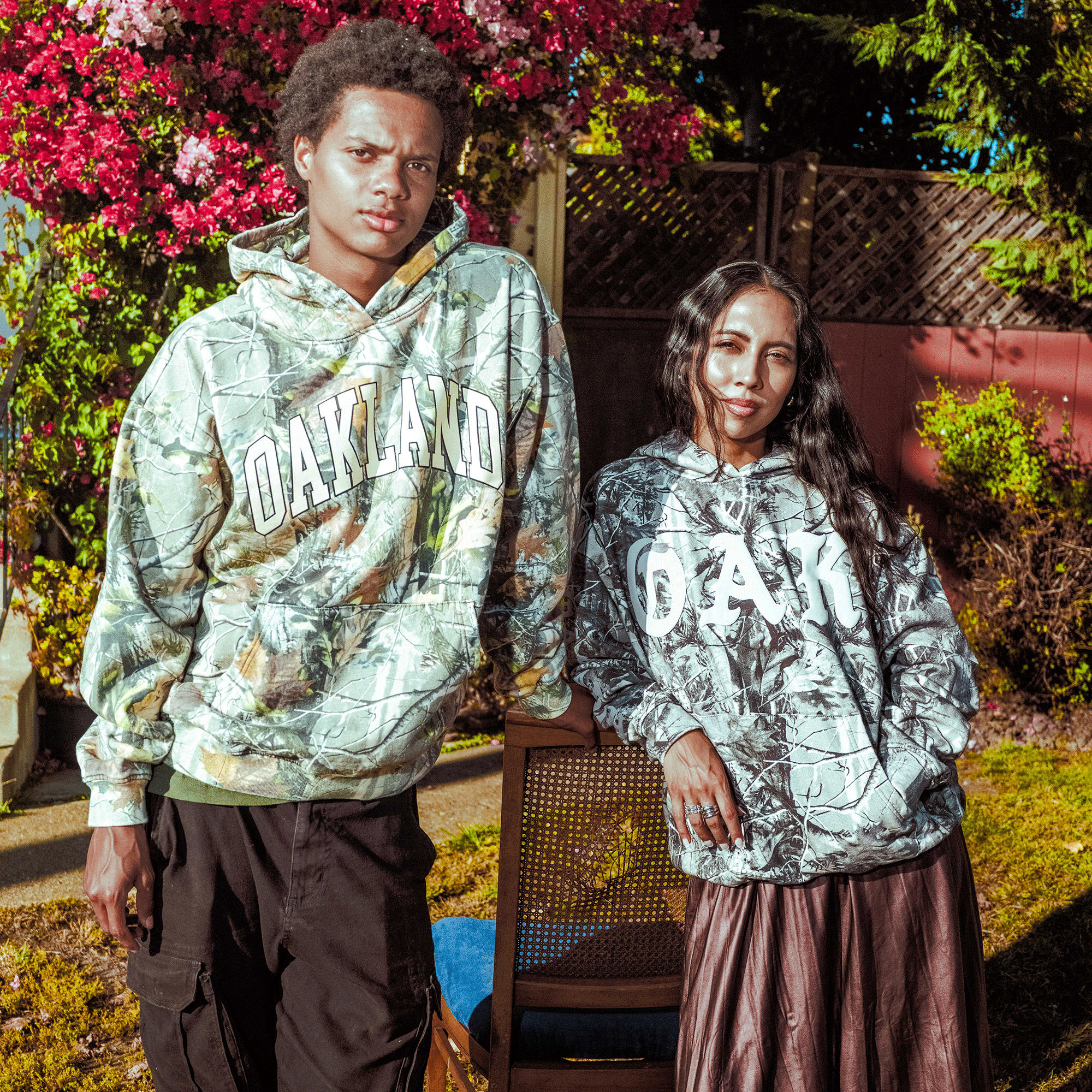 Two people stand outdoors in sunlit gardens, both wearing Oaklandish Cork Hoodies. The left wears cargo pants; the right, a long skirt. They lean on a chair amid blooming flowers and a wooden fence, highlighting these stylish hoodies.