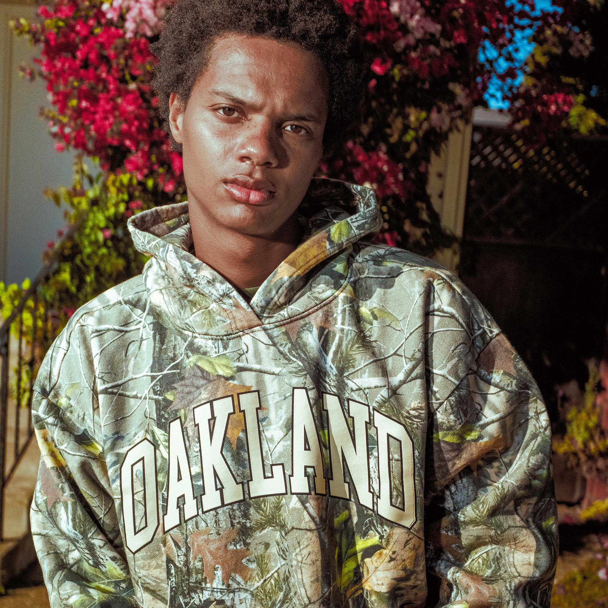 A young person stands outside in sunlight, wearing the Oaklandish Foliage Hoodie with "OAKLAND" in bold white letters and the Oaklandish tree logo. Surrounded by pink flowers and greenery, they have short curly hair and a serious expression.
