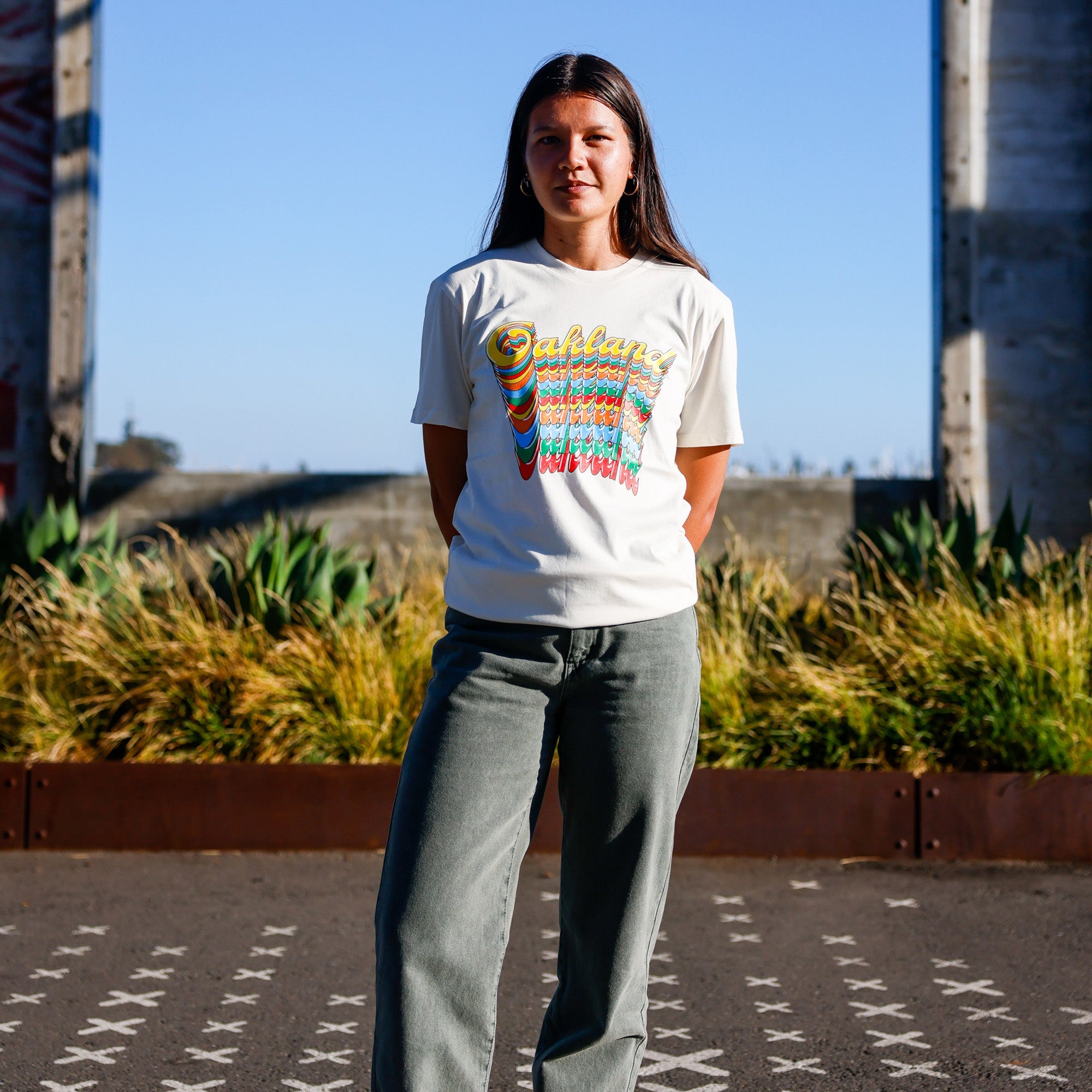 On a sunny day, a woman stands outdoors against a vibrant, rustic backdrop adorned with plants and tall concrete structures. Her straight, dark hair frames her face as she wears an "Oakland Roots SC Funk Tee" from Oakland Roots SC—a white T-shirt featuring multi-colored text that reads "Oakland Roots." She pairs this stylish tee with green pants and stands with her arms behind her back.