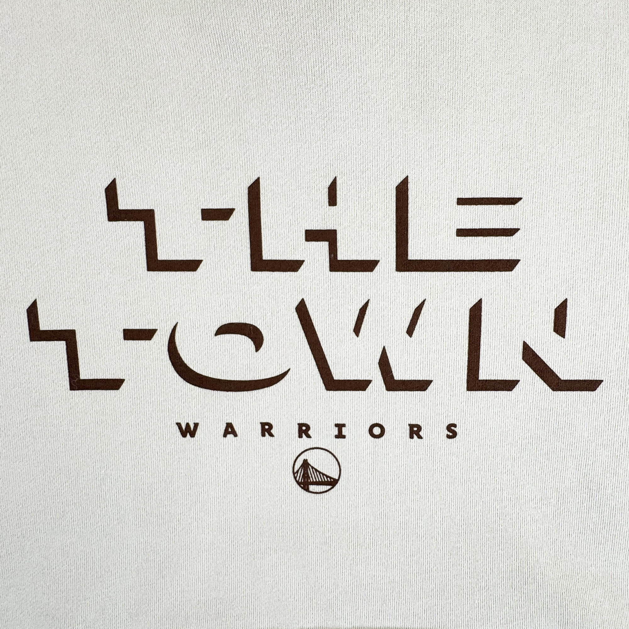 The GSW Ghost Block Letters Hoodie features white fabric with bold, angular brown “THE TOWN” text in a geometric font; beneath, “WARRIORS” appears above a stylized Oaklandish-inspired Golden State Warriors bridge logo.