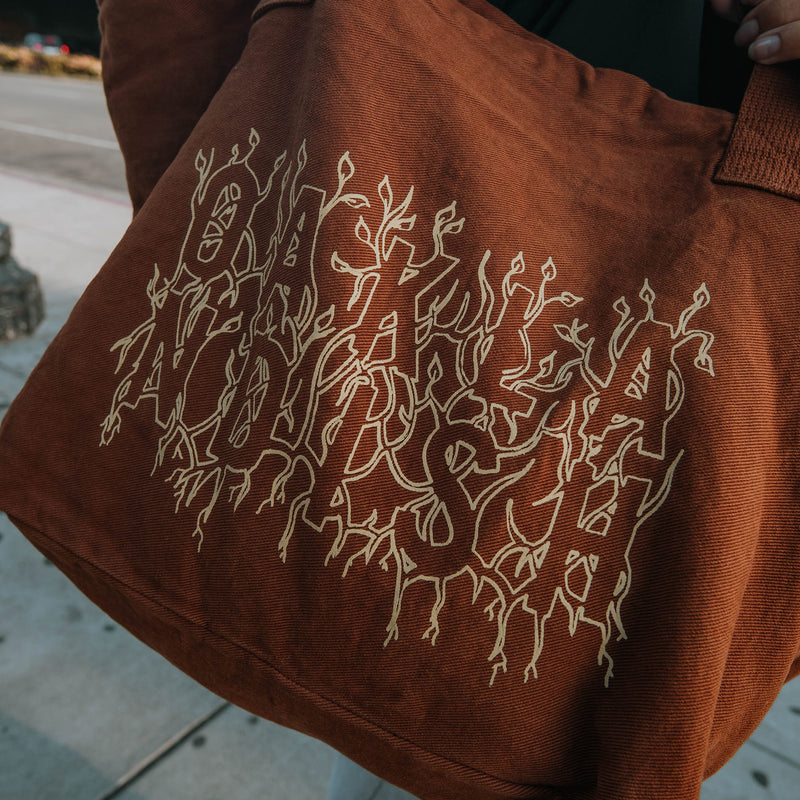 Close-up of creme ink and Oaklandish wordmark surrounded by stems and vines on Ginger tote bag in natural outdoor lighting.