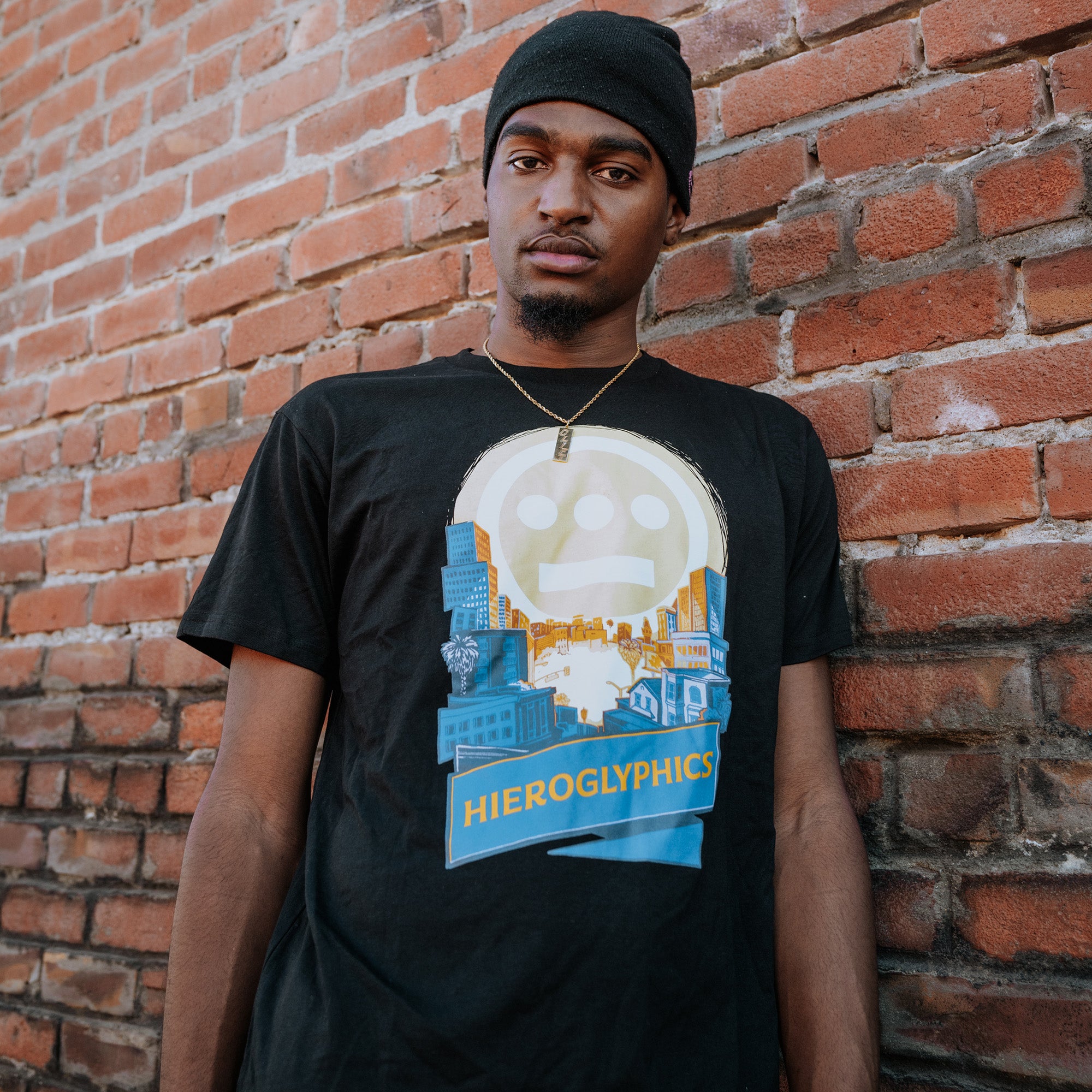Model leaning against brick wall wearing a black t-shirt with Hiero Valley, Hieroglyphics Hip Hop Graphic on the front chest.