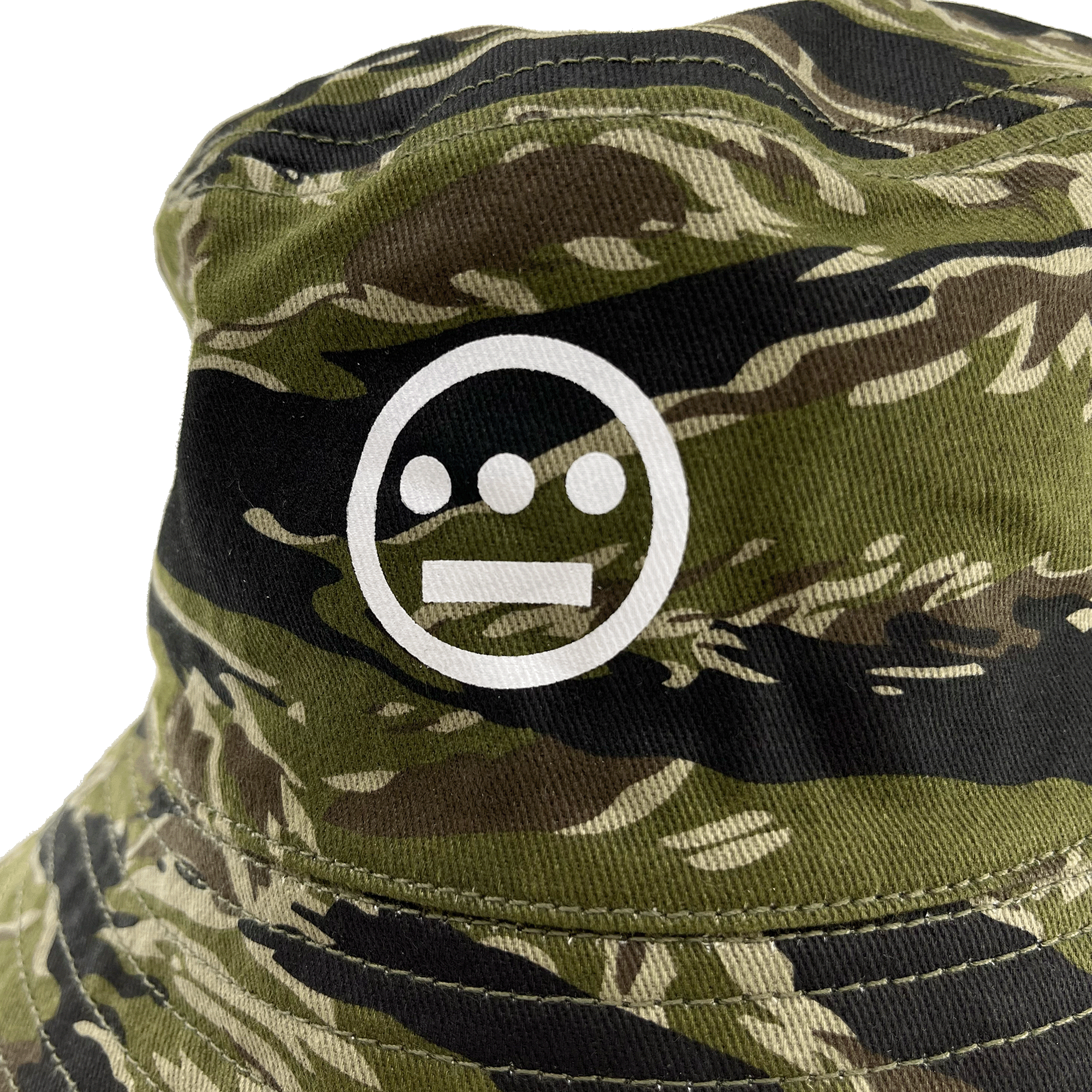 A close-up of the Hieroglyphics Hiero Reversible Camo Bucket hat showcases a white, minimalist face logo. This logo features three filled circles representing eyes and nose, and a horizontal line forming a simplistic mouth. The tiger camouflage pattern incorporates shades of green, brown, and beige.