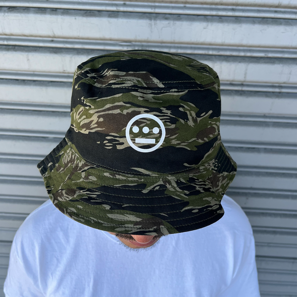 A man standing outdoors, face hidden, wearing a green camouflage bucket hat with a white Hieroglyphics Hip Hop Crew logo on the front crown. 