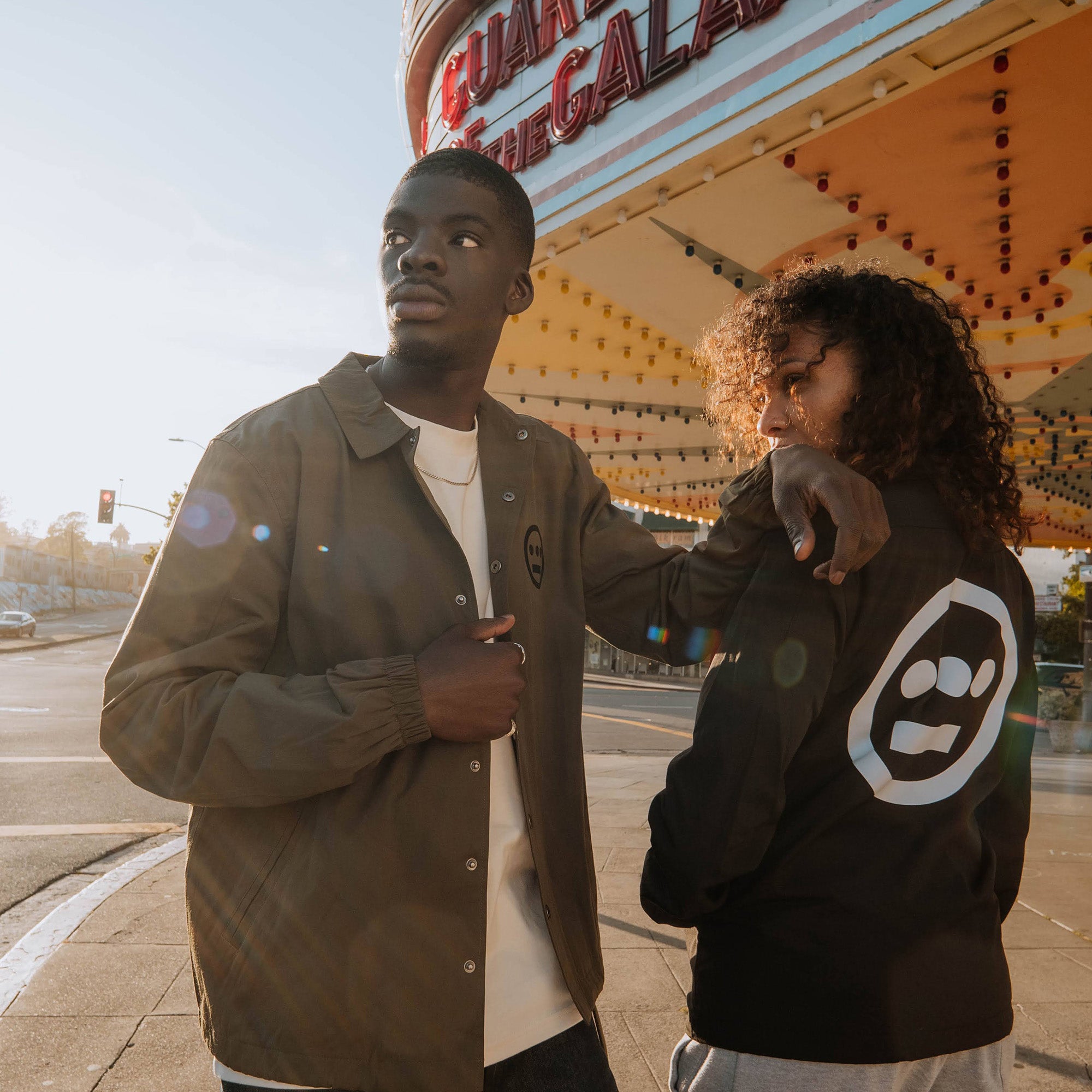 Male and female model near movie marquee with male wearing olive jacket with black left chest hiero logo and female model wearing black jacket with large hiero logo on back.