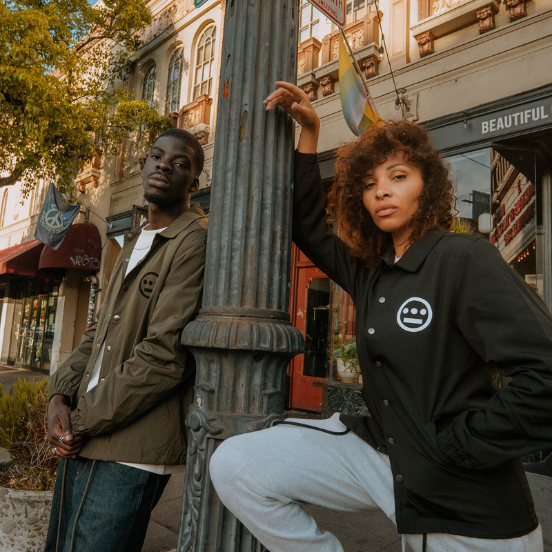 Male and female model leaning against lightpost with male wearing olive jacket with black left chest hiero logo and female model wearing black jacket with large hiero logo left chest in white.