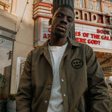 male model leabing against lightpost in front of move marquee wearing olive jacket unbuttoned with hiero third eye logo in black on left chest, olive jacket.