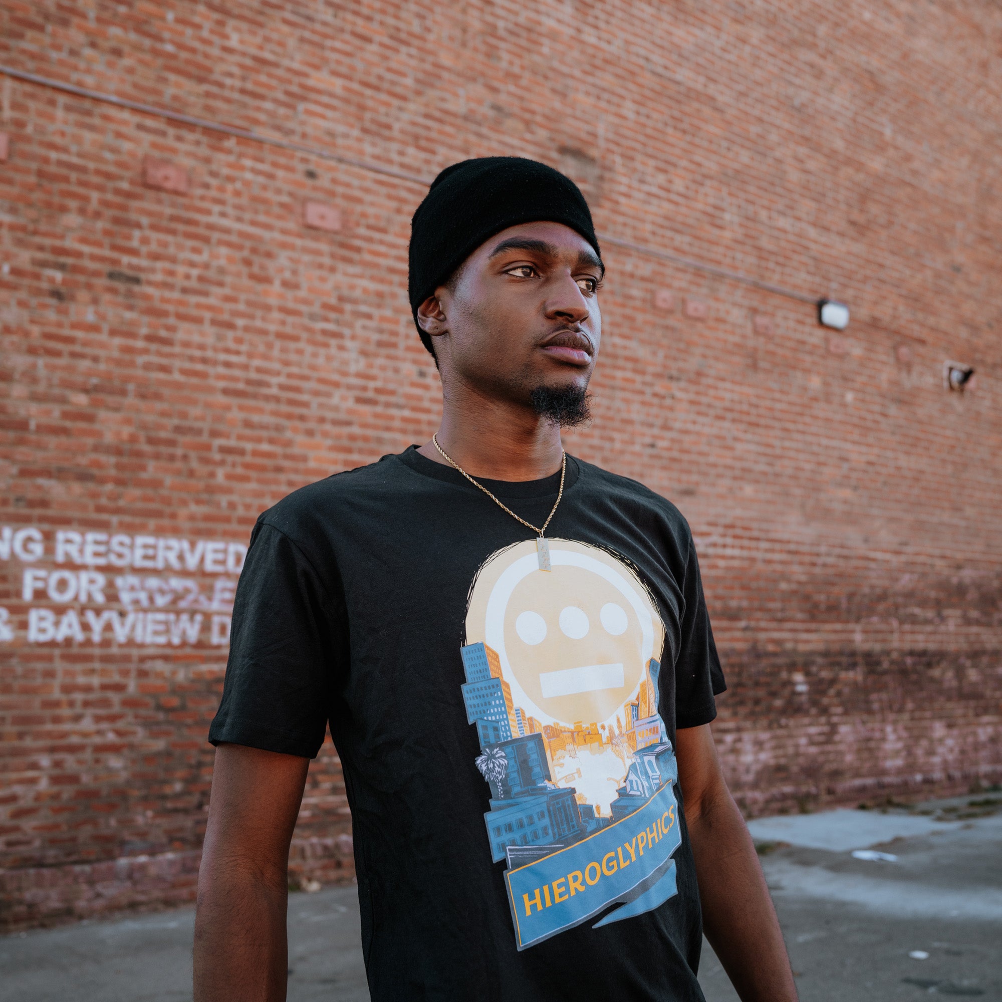 A man wearing a black beanie, gold necklace, and Hieroglyphics' Hiero Valley Tee stands against a red-brick wall. The 100% cotton T-shirt features a stylized face and cityscape design. The wall has faded white text, and the ground is partially visible.