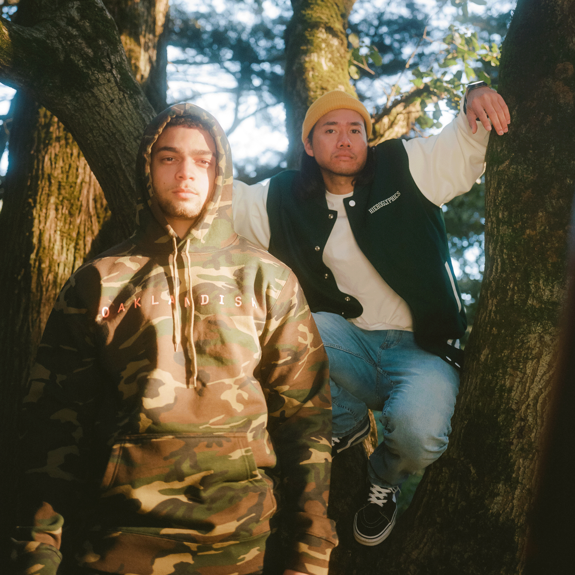 Two men outdoors, sitting in and standing by a tree in Oaklandish lifestyle apparel. 
