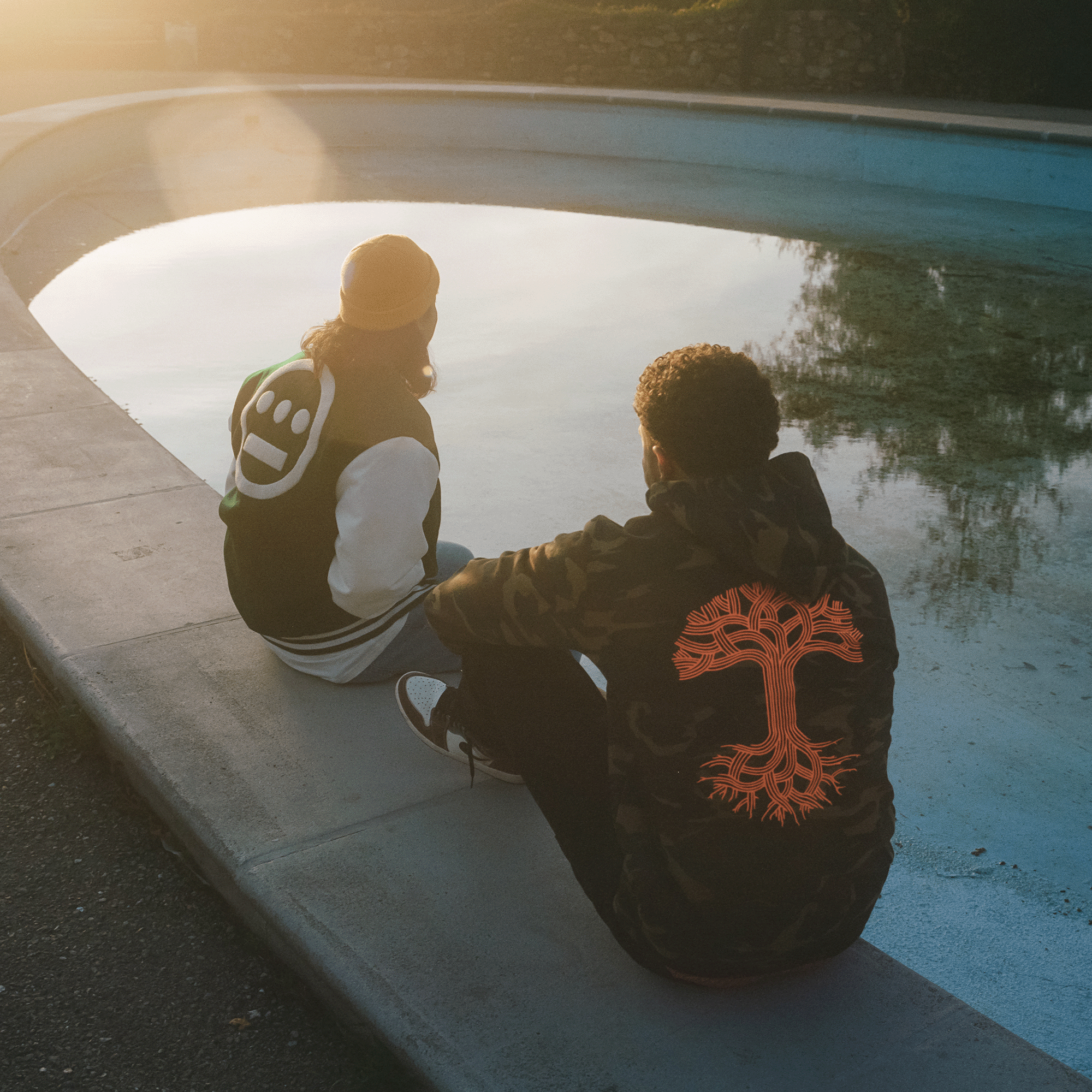 Back view of a model sitting by pool wearing camo pullover hoodie sweatshirt with a large orange Oaklandish tree logo in the center.