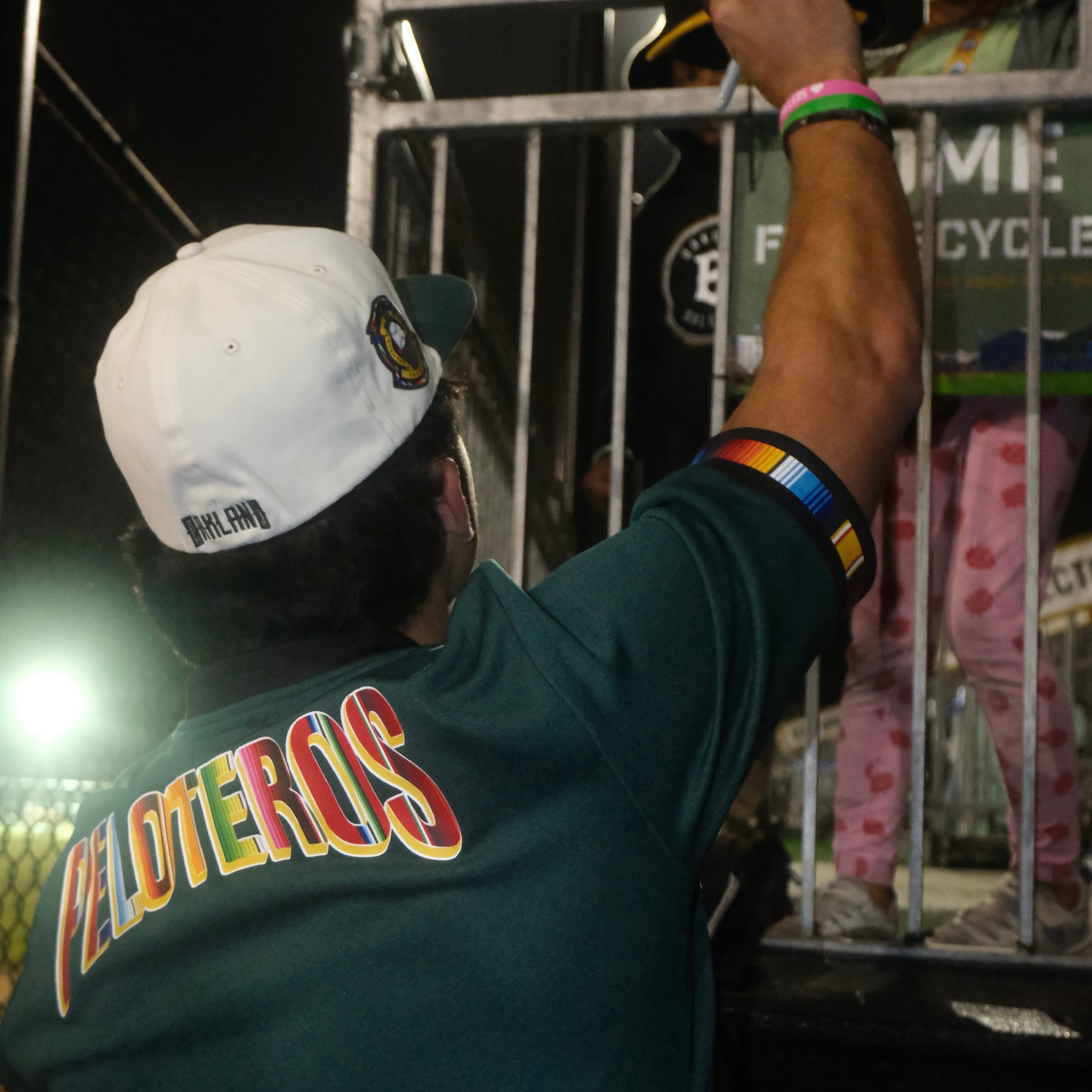 A person in a green jersey labeled "PELOTEROS" reaches up toward a child in pink heart pajamas behind a metal railing. The person—perhaps an Oakland Ballers supporter—wears an Oakland Peloteros Hat by Oakland Ballers, along with rainbow armbands; the nighttime scene is illuminated by bright lights in the background.